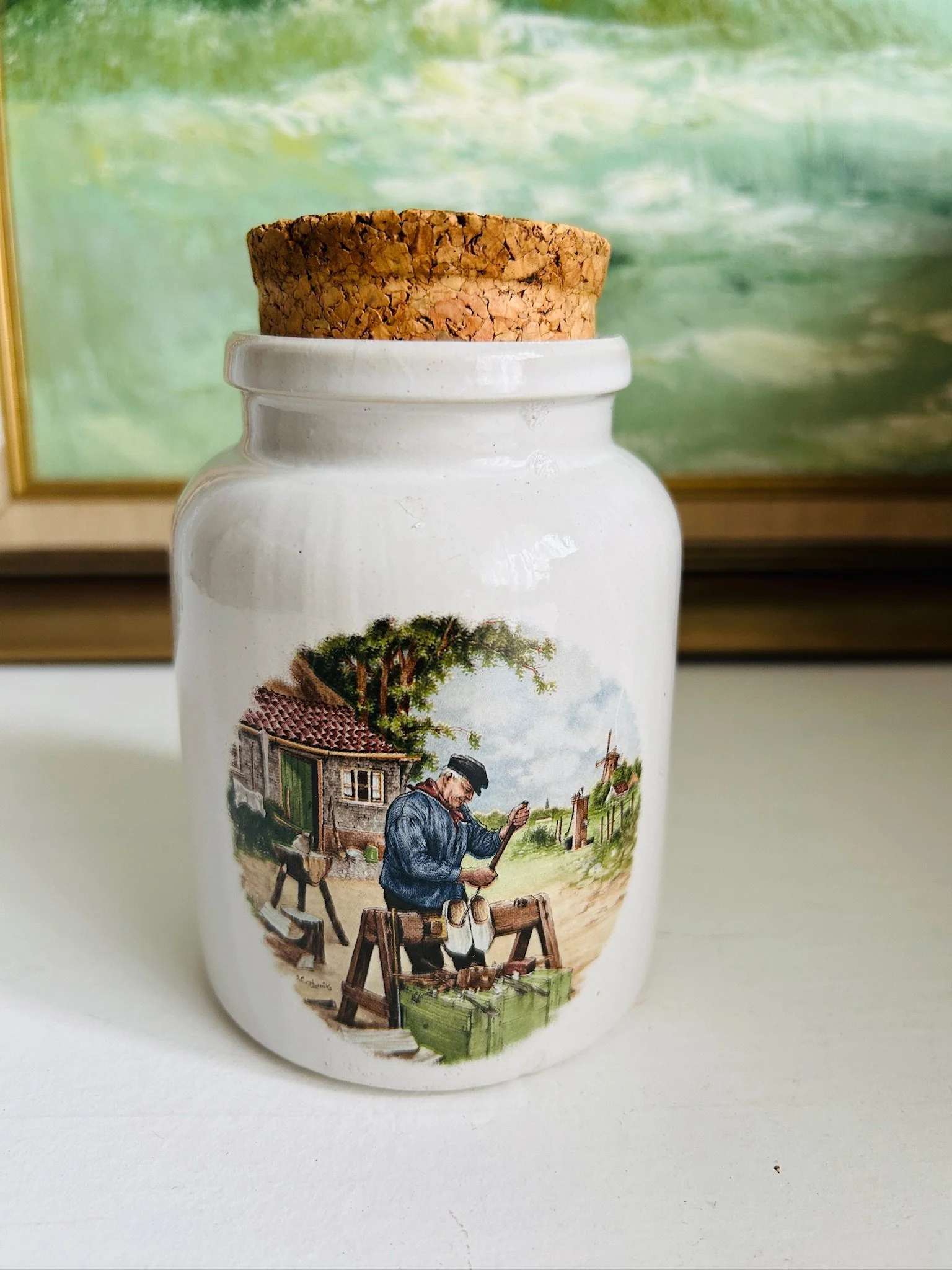Single ceramic pot with cork lid showing the cobbler village scene, painted in soft earthy tones on a cream background.