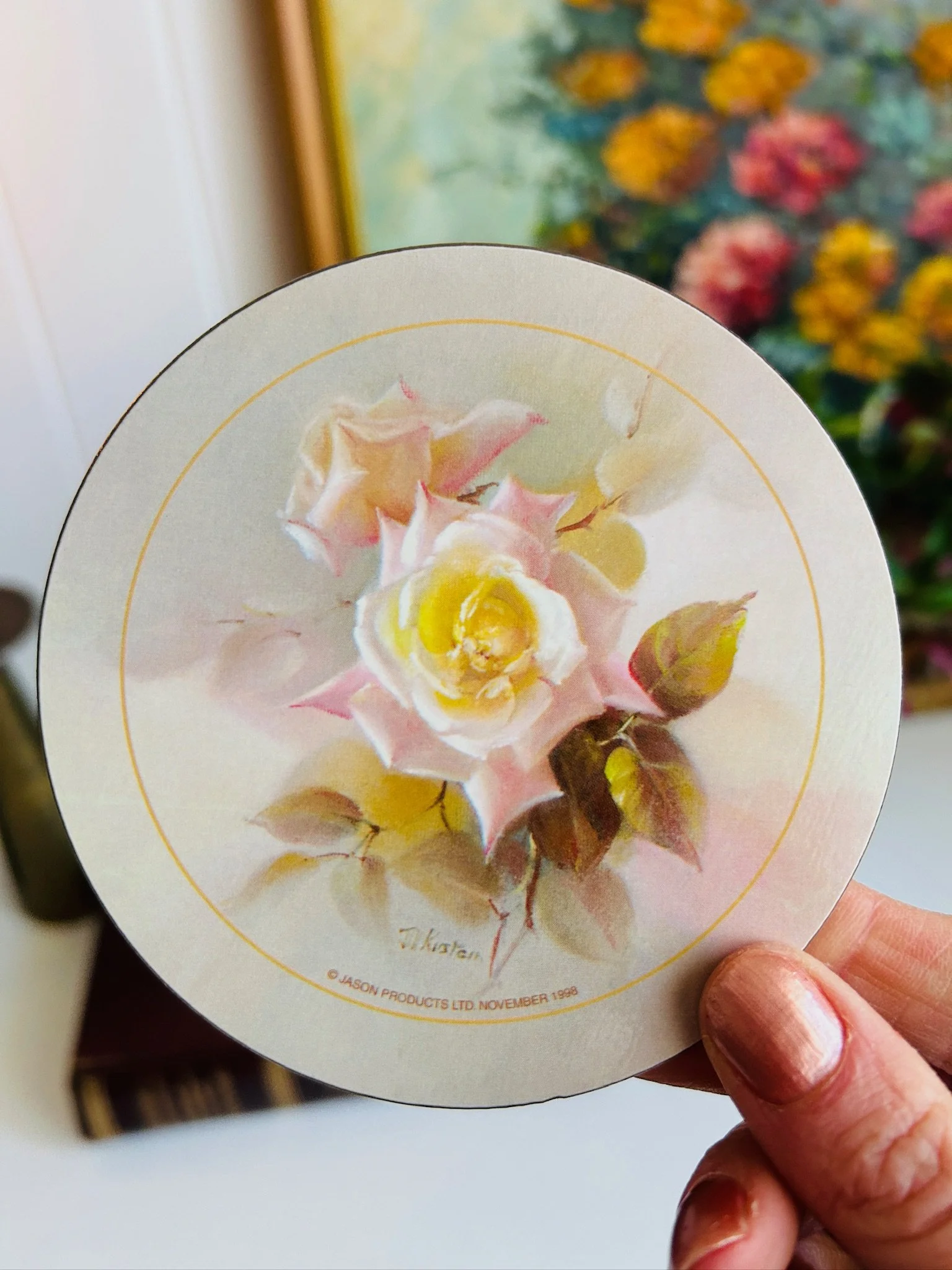 Hand holding a 24 cm round Jason placemat with a pale pink and yellow rose painted by Jill Kirstein