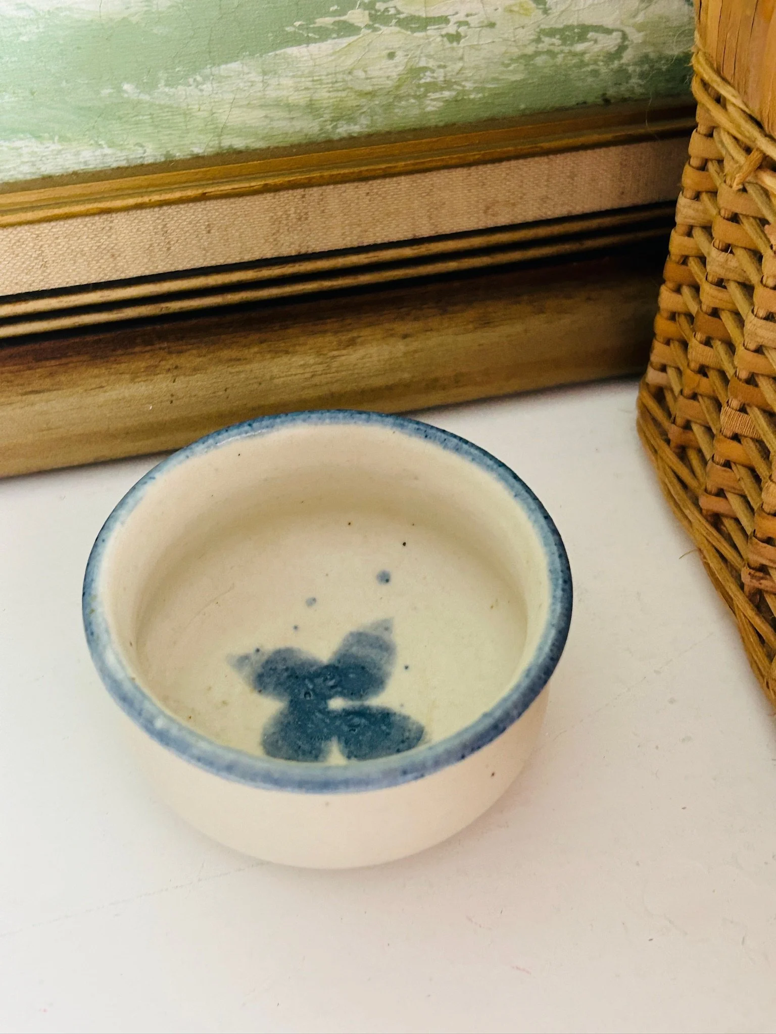 Small artisan ceramic bowl with beige glaze, blue rim, and blue floral centre detail styled beside a wicker basket and framed artwork