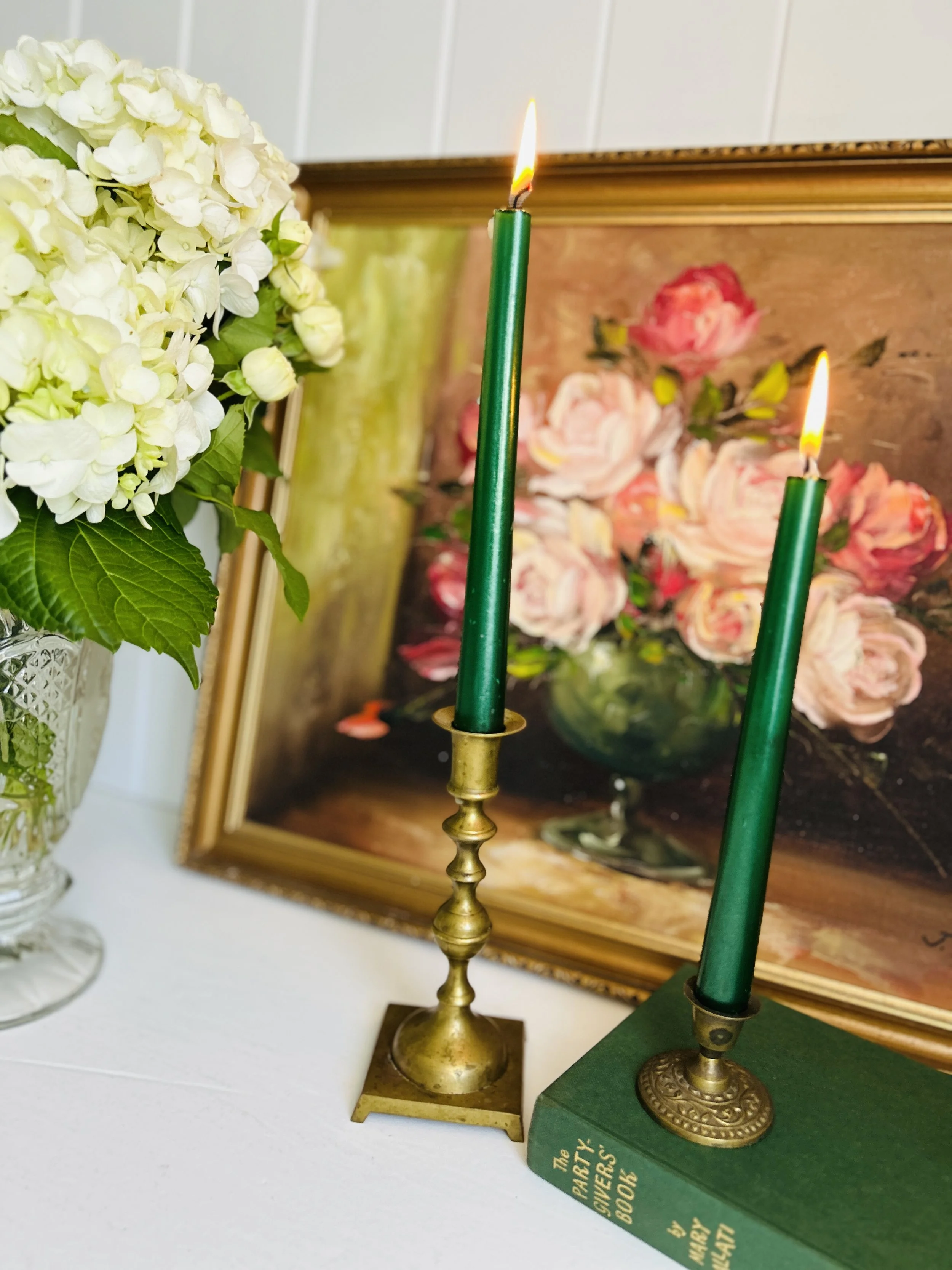 A soft-focus scene with pale green taper candles glowing in small crystal candleholders beside a cream vintage vase decorated with hand-painted pink and white flowers and filled with blue hydrangeas, with stacked green and navy books and a floral pai
