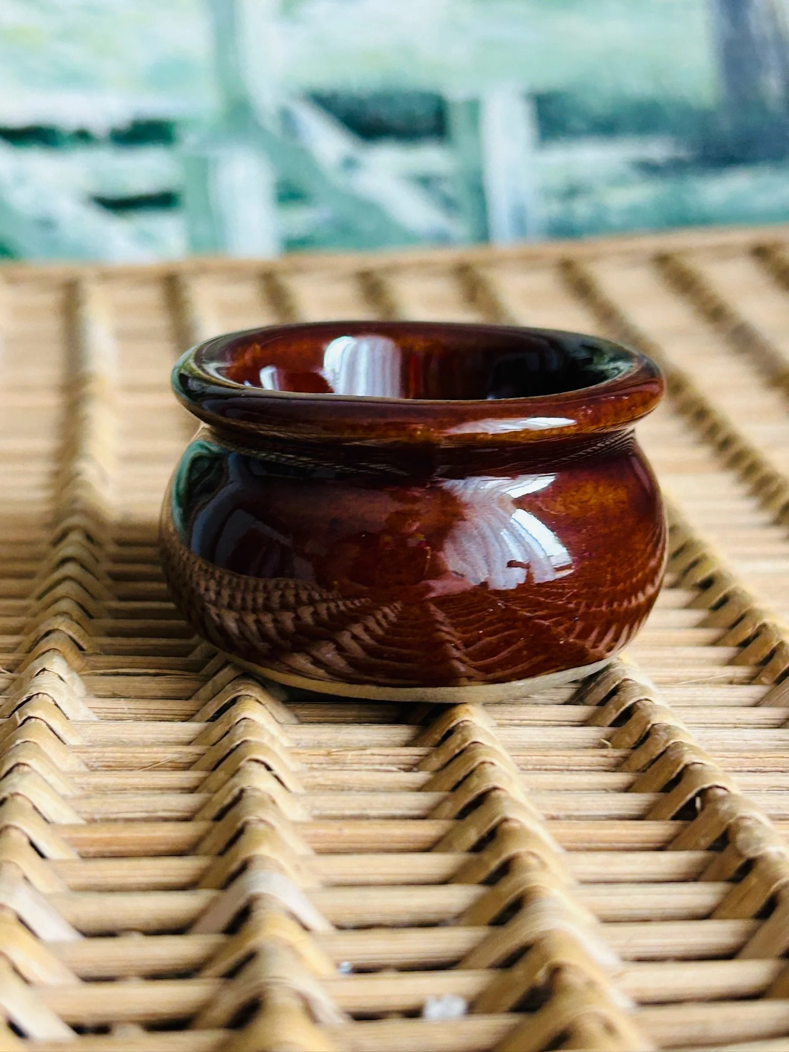 Front angled view of a small Moira Pottery salt cellar with glossy brown glaze resting on a woven wicker surface