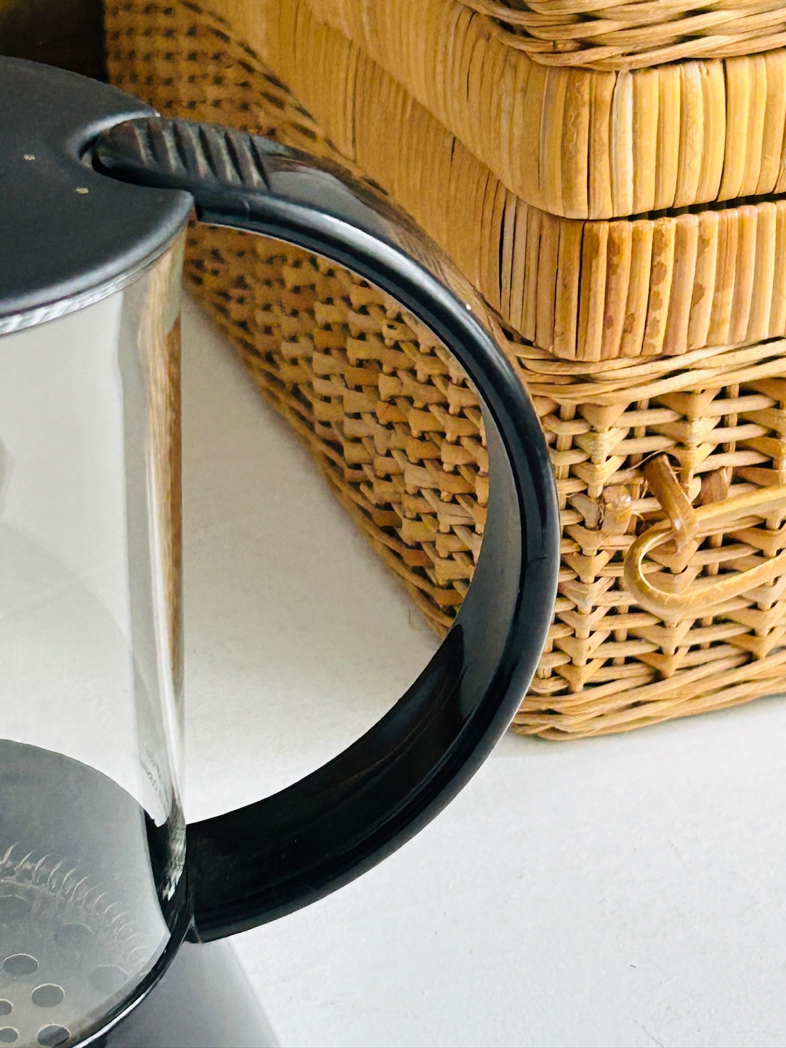 French press held in hand, showing the full profile with black handle, lid, and clear glass body against a floral painting background.