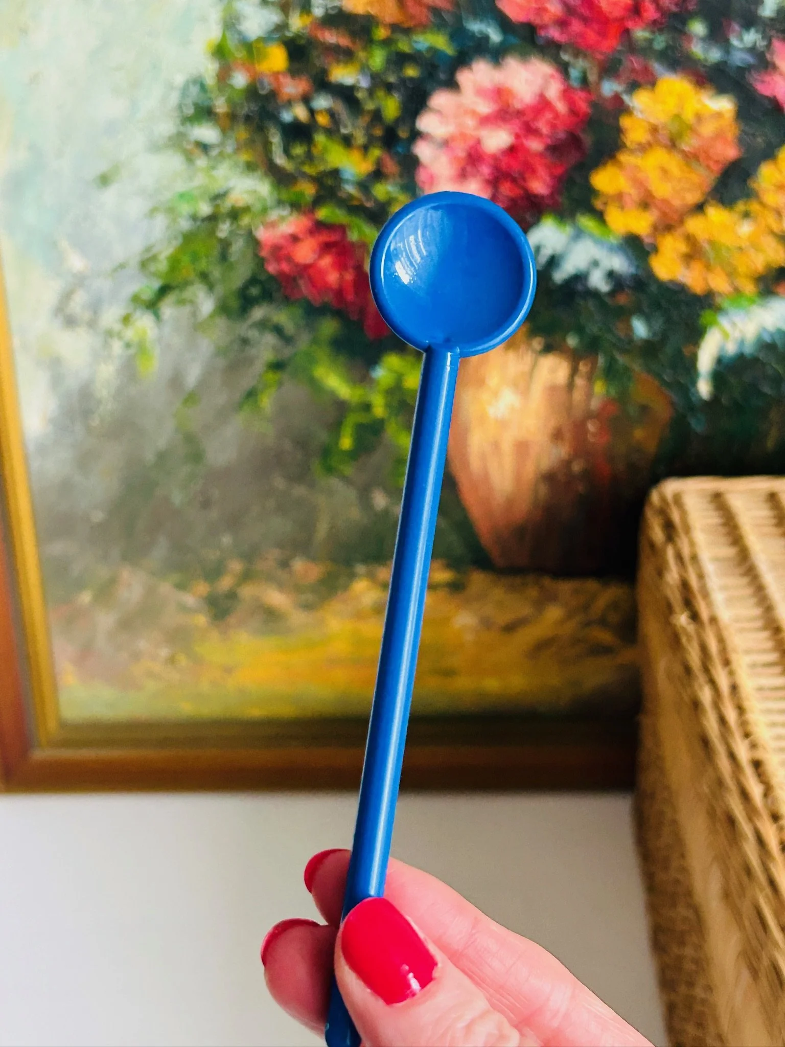 Single Bodum blue stir spoon shown upright in front of floral artwork and basket background