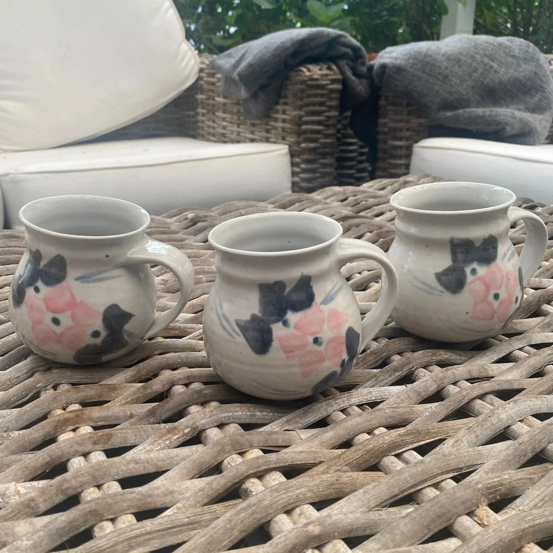 Set of three hand-painted ceramic mugs on wicker surface, showing full shape and floral design