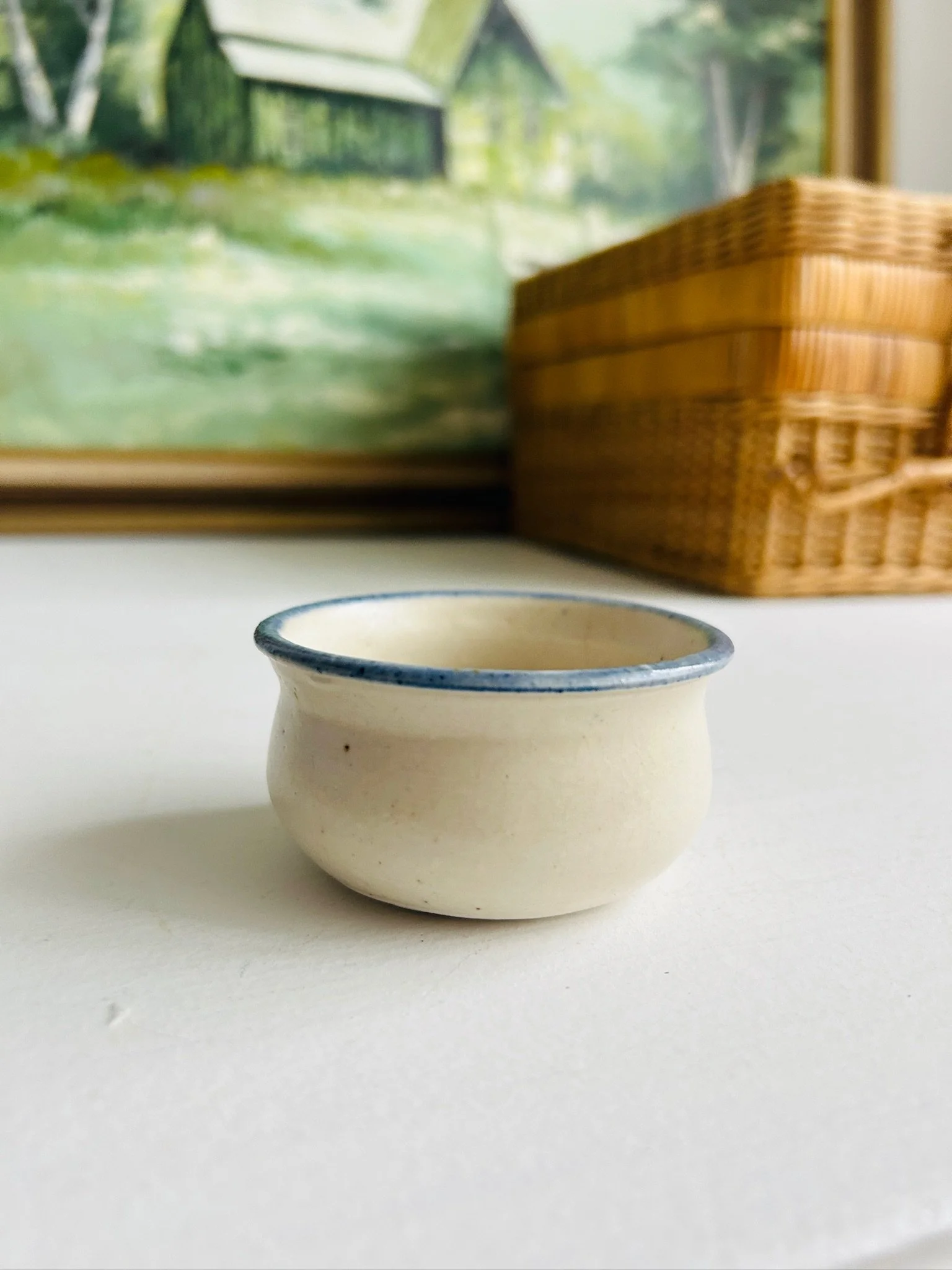 Full profile of the petite pottery bowl on a white surface with basket and landscape artwork in the background