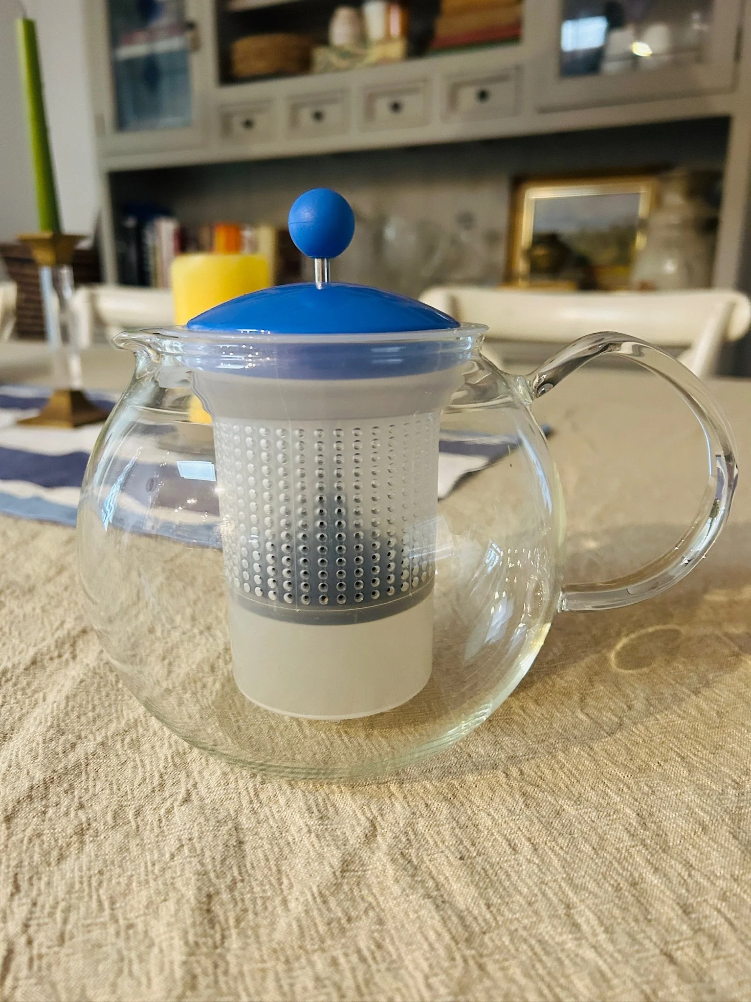 Side view of the Bodum Assam teapot on a table, showing the rounded glass body, clear handle, blue lid, and internal infuser.
