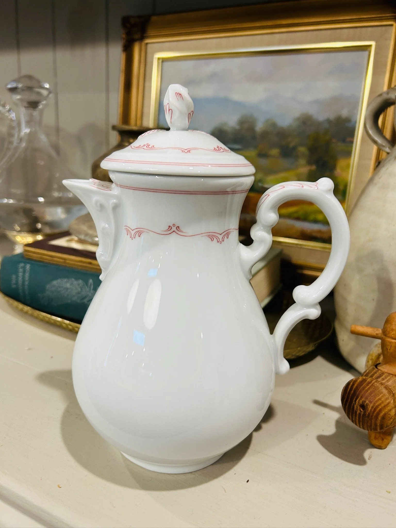 Three-quarter view of the porcelain coffee pot styled on a shelf, highlighting the curved handle, domed lid, and soft red floral motif.