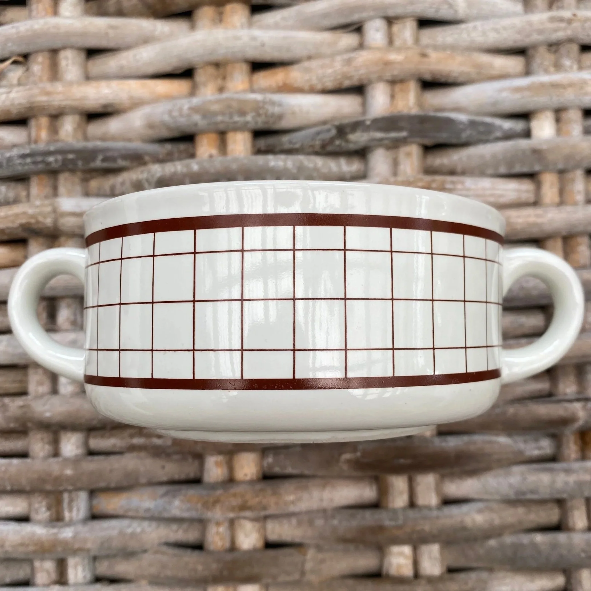 Brown grid pattern soup mug with twin handles on woven surface