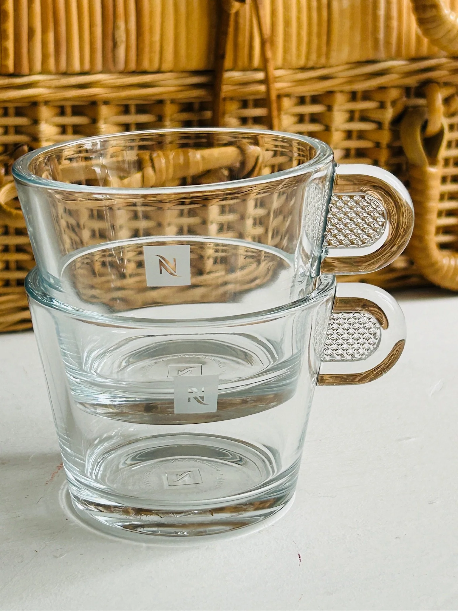 Two stacked clear glass Nespresso espresso cups with etched N logos and textured handles shown against a woven basket background