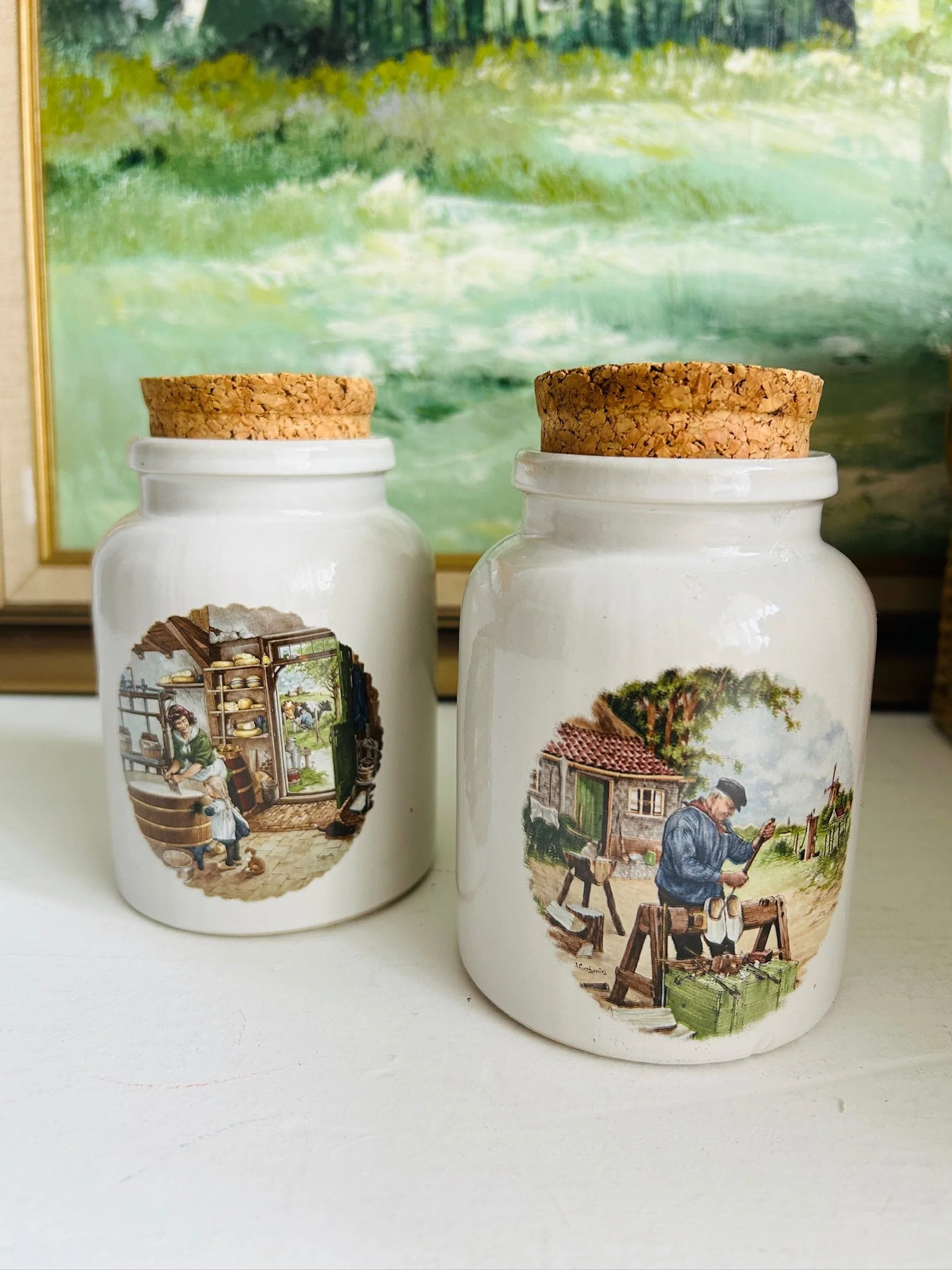 Full set of two Dutch ceramic pots side by side, one with a cheese shop scene and one with a cobbler scene, against a painted backdrop.