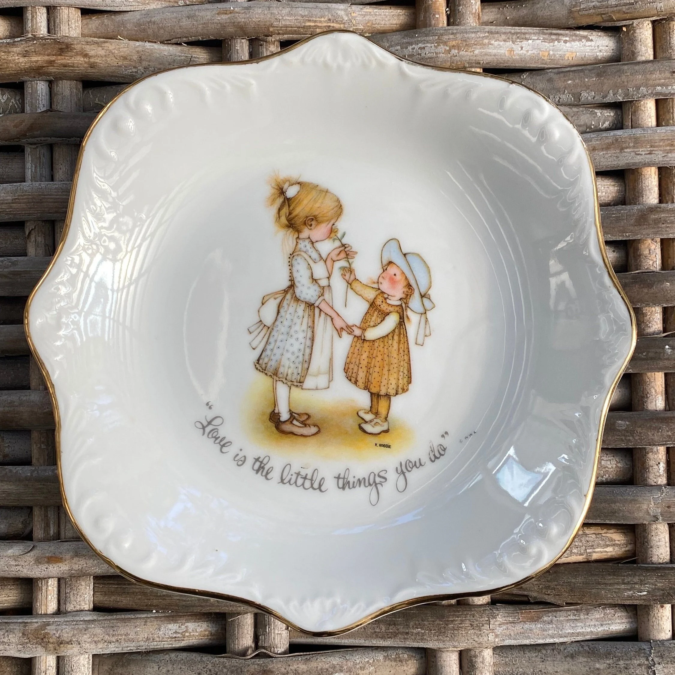 Front of Holly Hobbie 1973 porcelain plate featuring two children and the quote 'Love is the little things you do...', on a wicker background