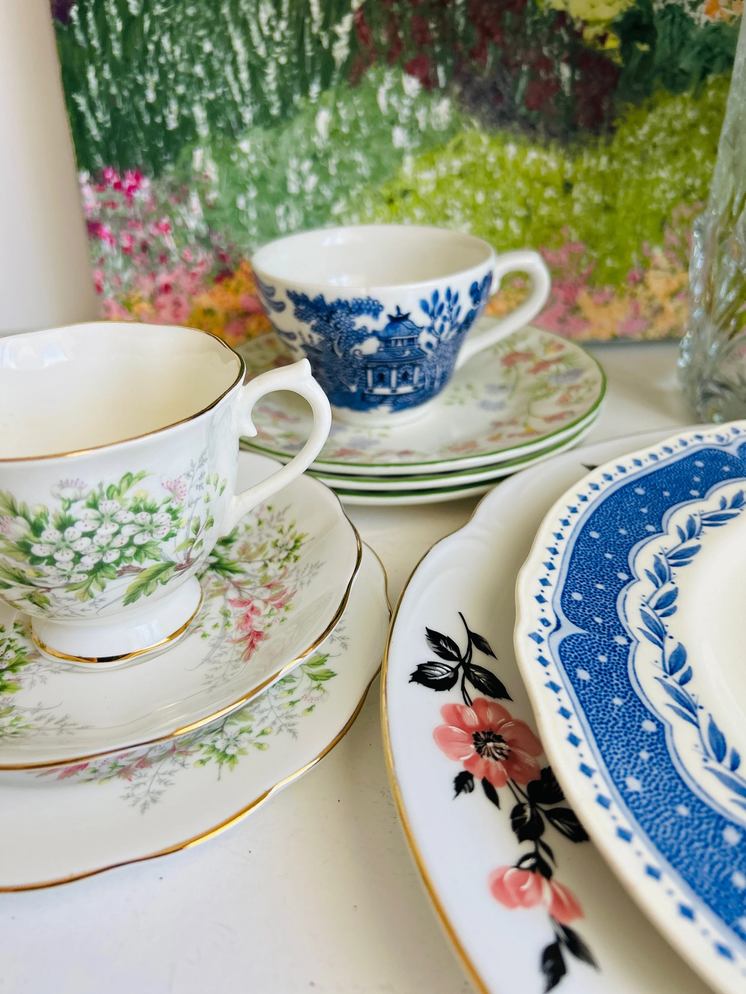 Close-up of vintage porcelain teacups and saucers with floral and scenic patterns, arranged on a white table with a colorful floral background.