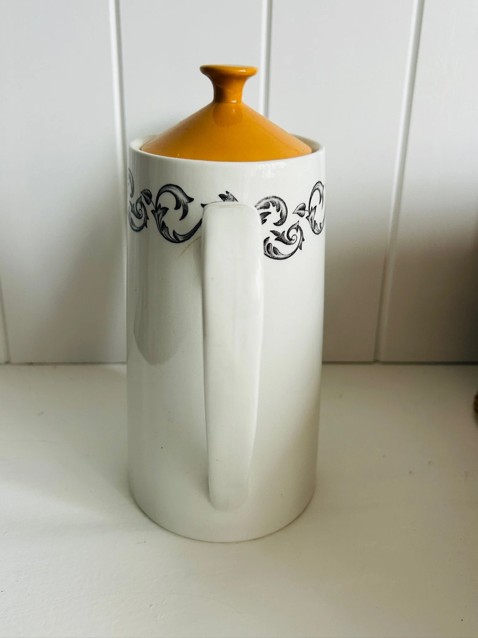 Rear view of the coffee pot showing the full handle profile, orange lid finial, and black scrollwork pattern wrapping around the upper body.