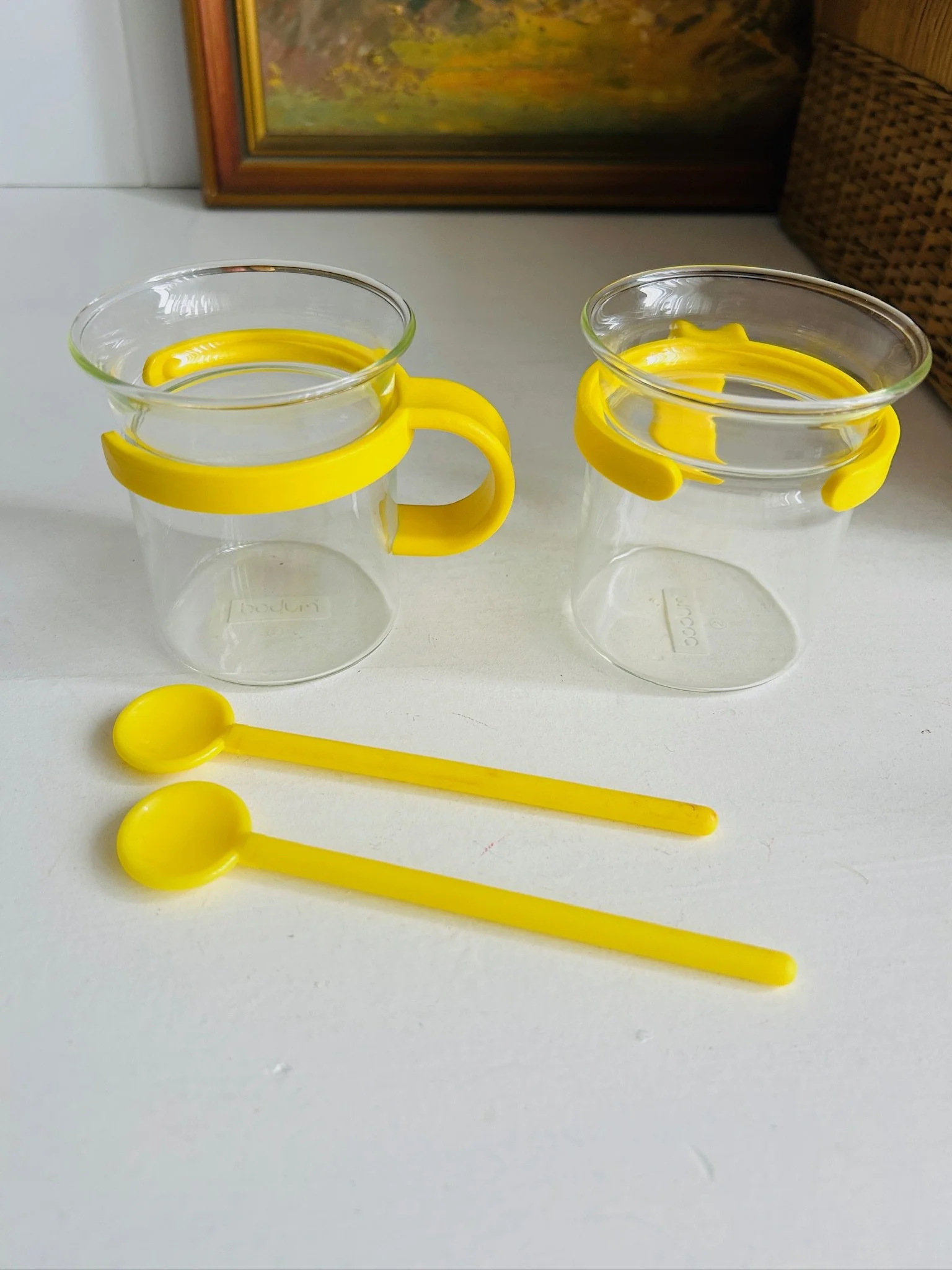 Pair of Bodum Picard cups with yellow holders and matching yellow spoons arranged on a white shelf.