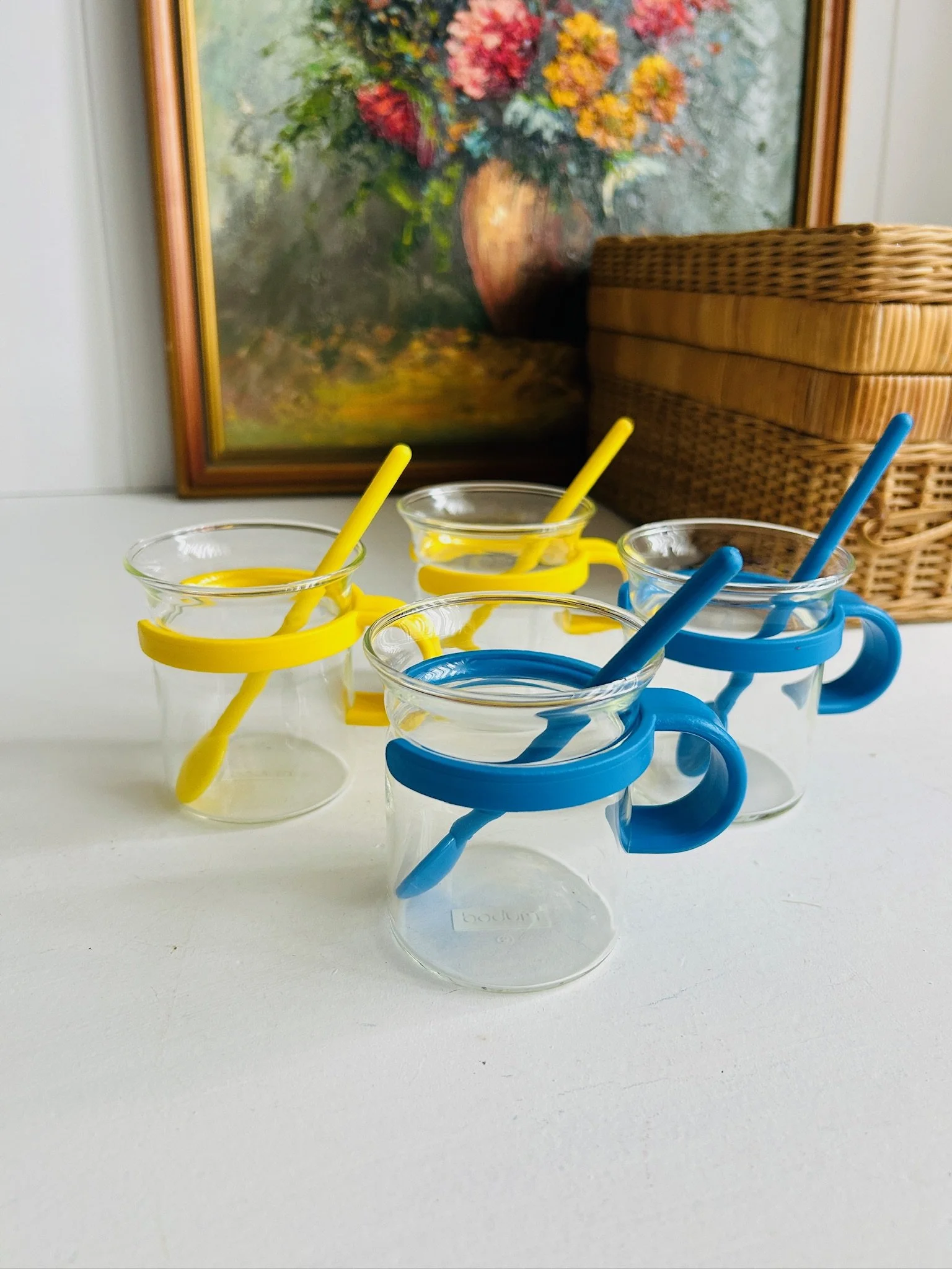 Group of four vintage Bodum Picard glass cups with yellow and blue plastic holders and matching spoons, arranged together on a white surface.