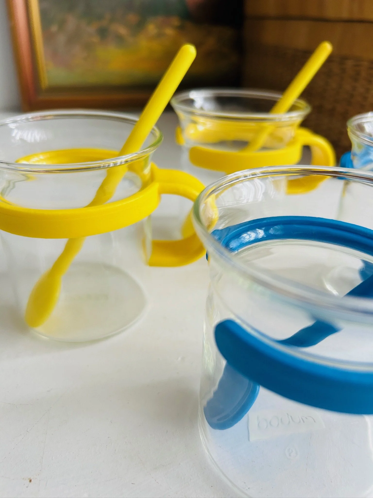 Close-up of Bodum Picard cups showing the clear glass, bright yellow holder, blue holder, and matching long-handled spoons.