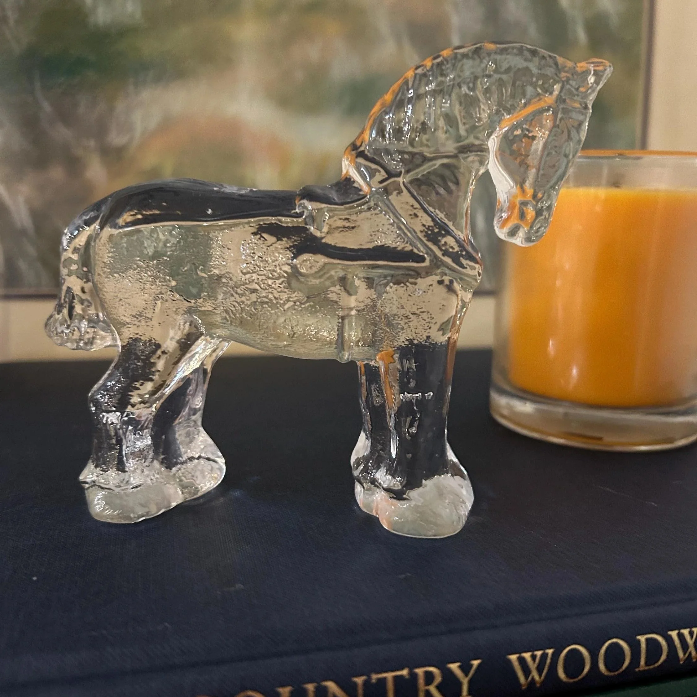 Side view of Kosta Boda glass horse figurine on a book beside a candle