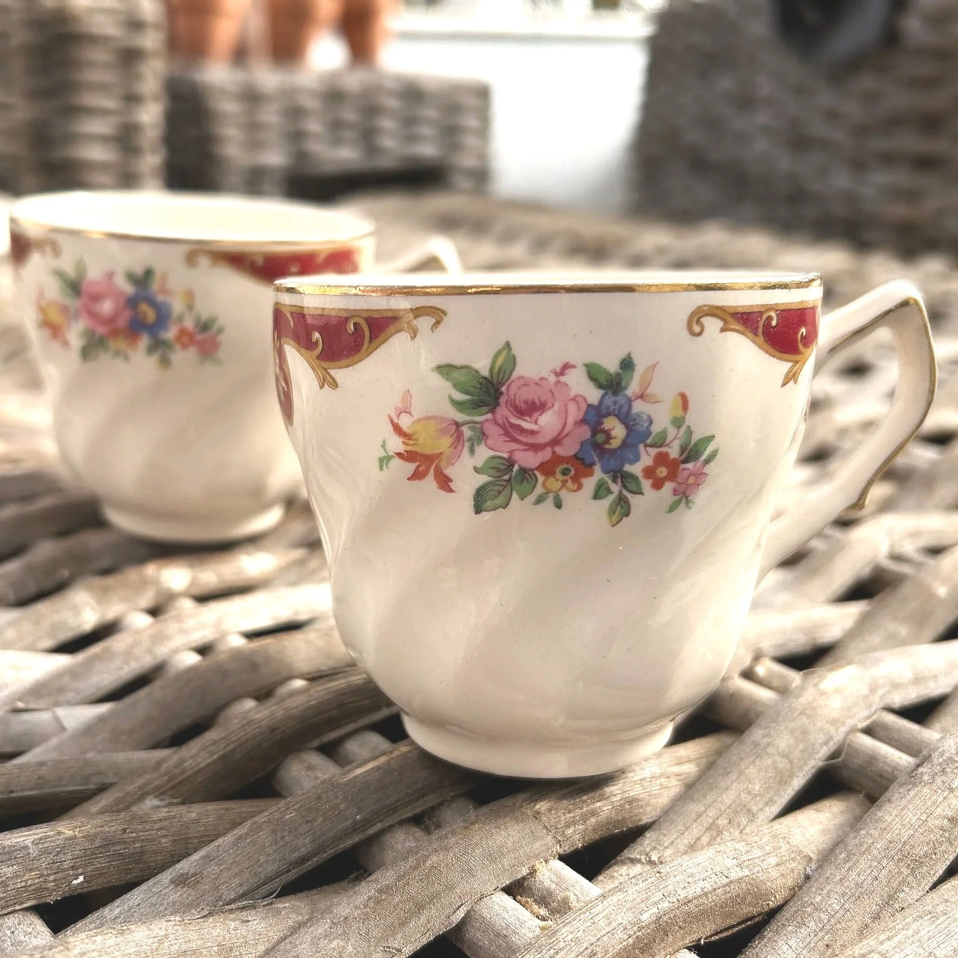 : Pair of English teacups with red and gold trim and floral design on wicker surface