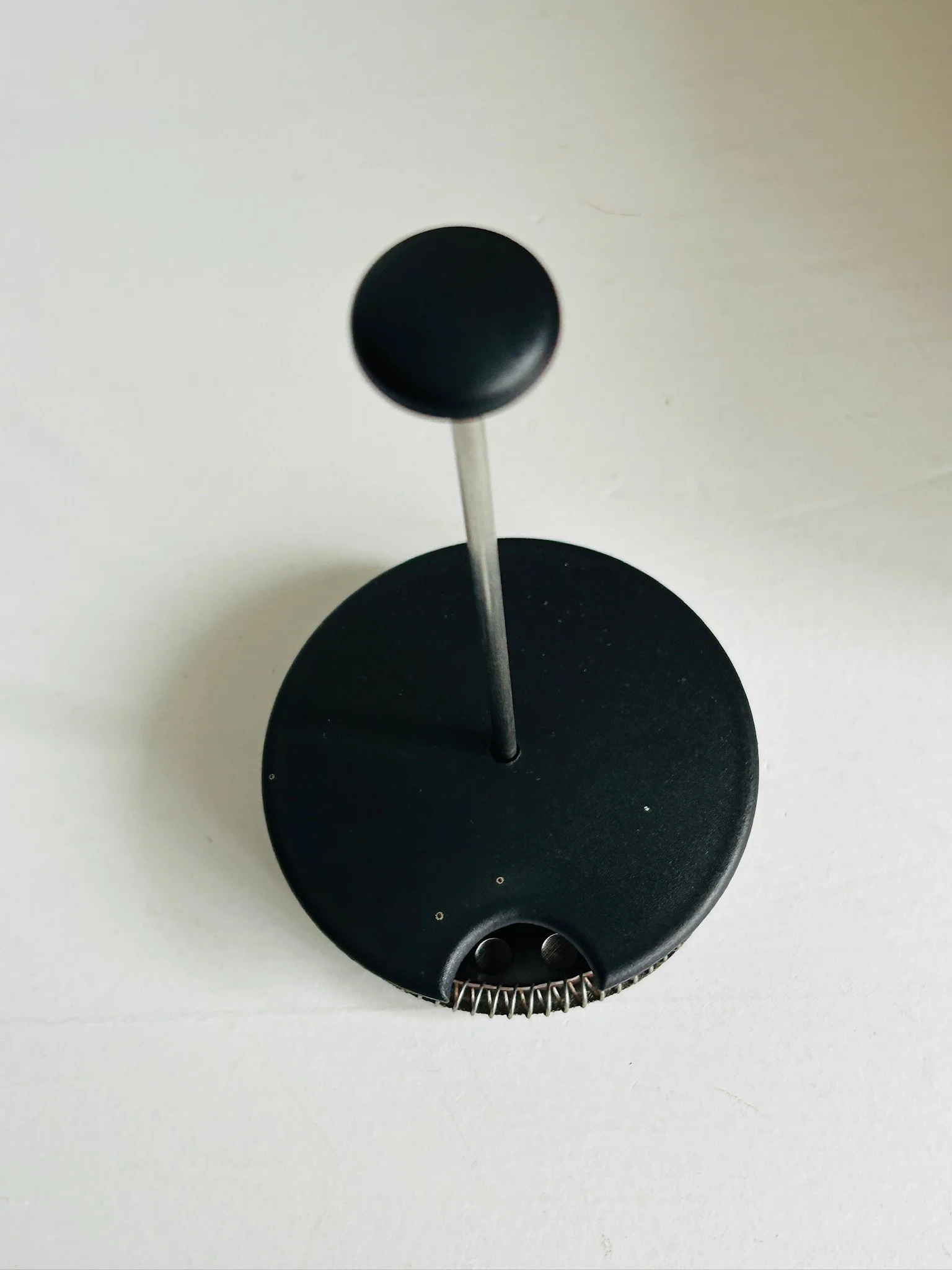 Top view of the black lid and plunger rod removed from the French press, showing the round knob and central metal shaft.