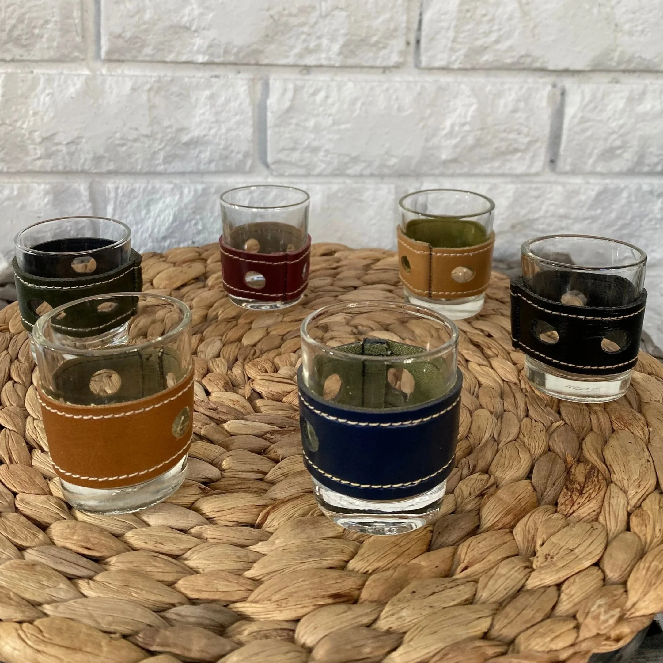 Full set of 6 leather-banded shot glasses displayed on a woven placemat with white brick background