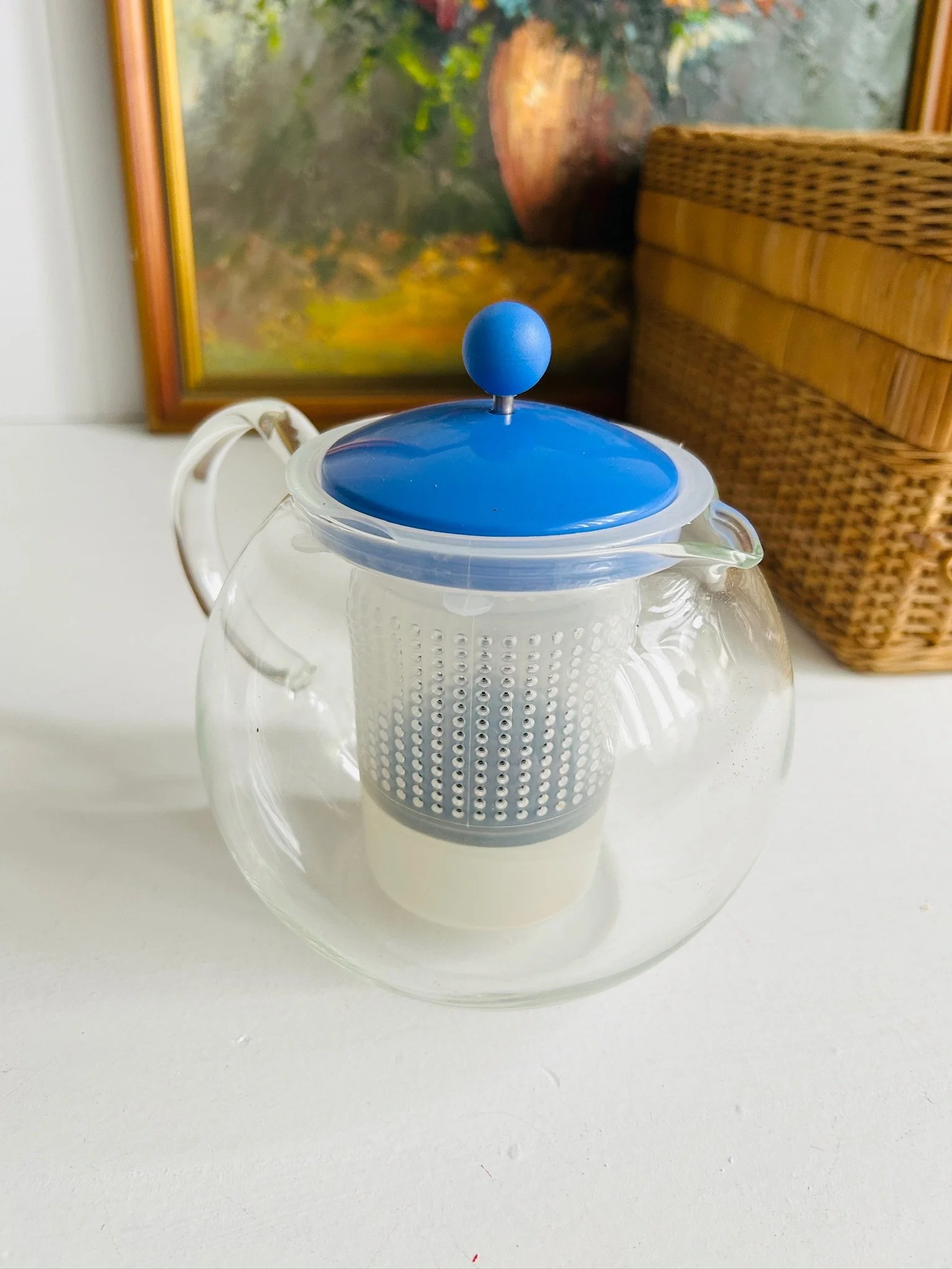 Three-quarter view of the assembled Bodum Assam tea press with blue lid and clear glass handle against a wicker backdrop.