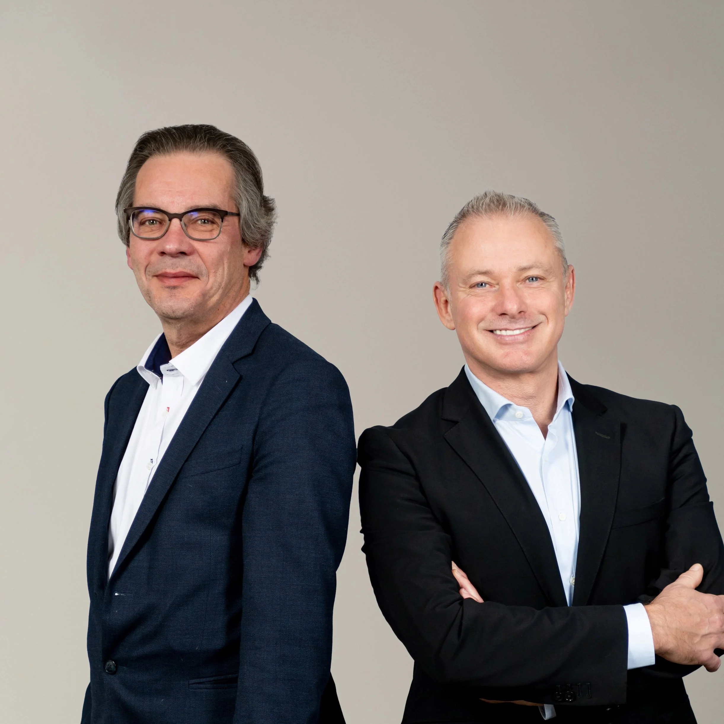 Two men standing side by side, dressed in business suits, smiling with arms crossed against a plain, light-colored background.