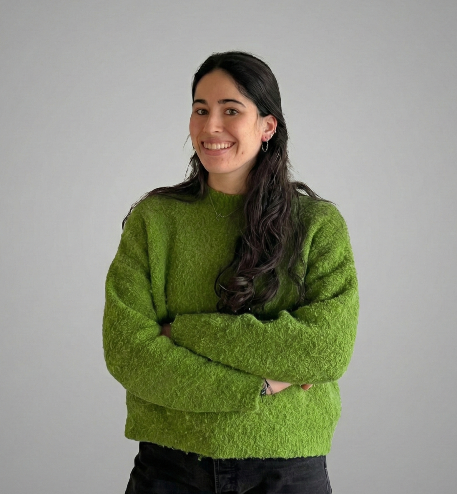A confident young woman with dark brown hair and fair skin, wearing a gray blazer and white top, standing with her arms crossed against a light gray background.