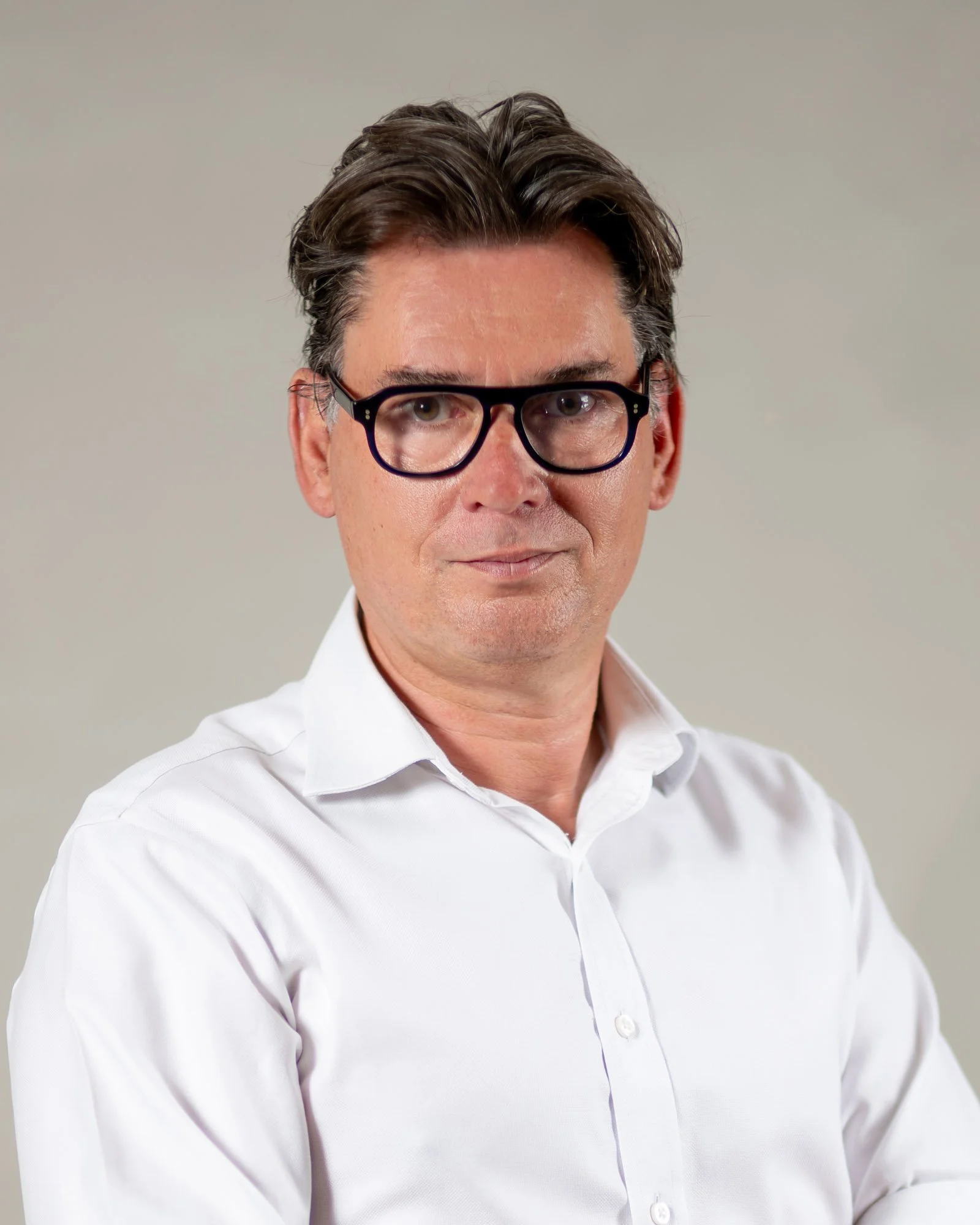 Portrait of a man with dark hair, wearing large black glasses, a white dress shirt, standing against a neutral background.