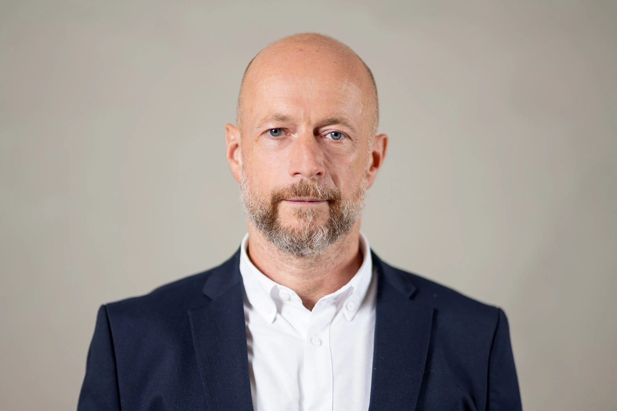 Portrait of a middle-aged man with a bald head and gray beard wearing a white dress shirt and navy blazer against a plain neutral background.
