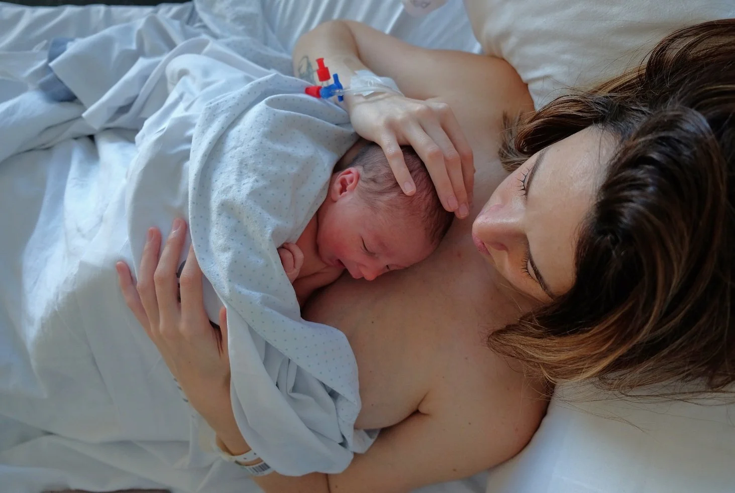 Mother with newborn baby in bed, skin-to-skin contact, both wrapped in blanket.