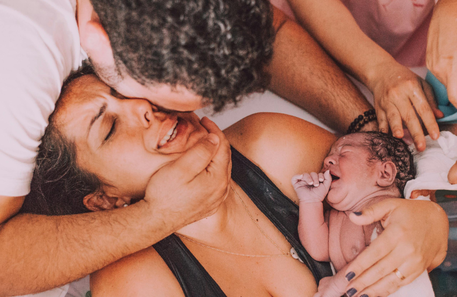 A new mother holding her newborn baby with emotional expressions while a partner embraces her from the side.