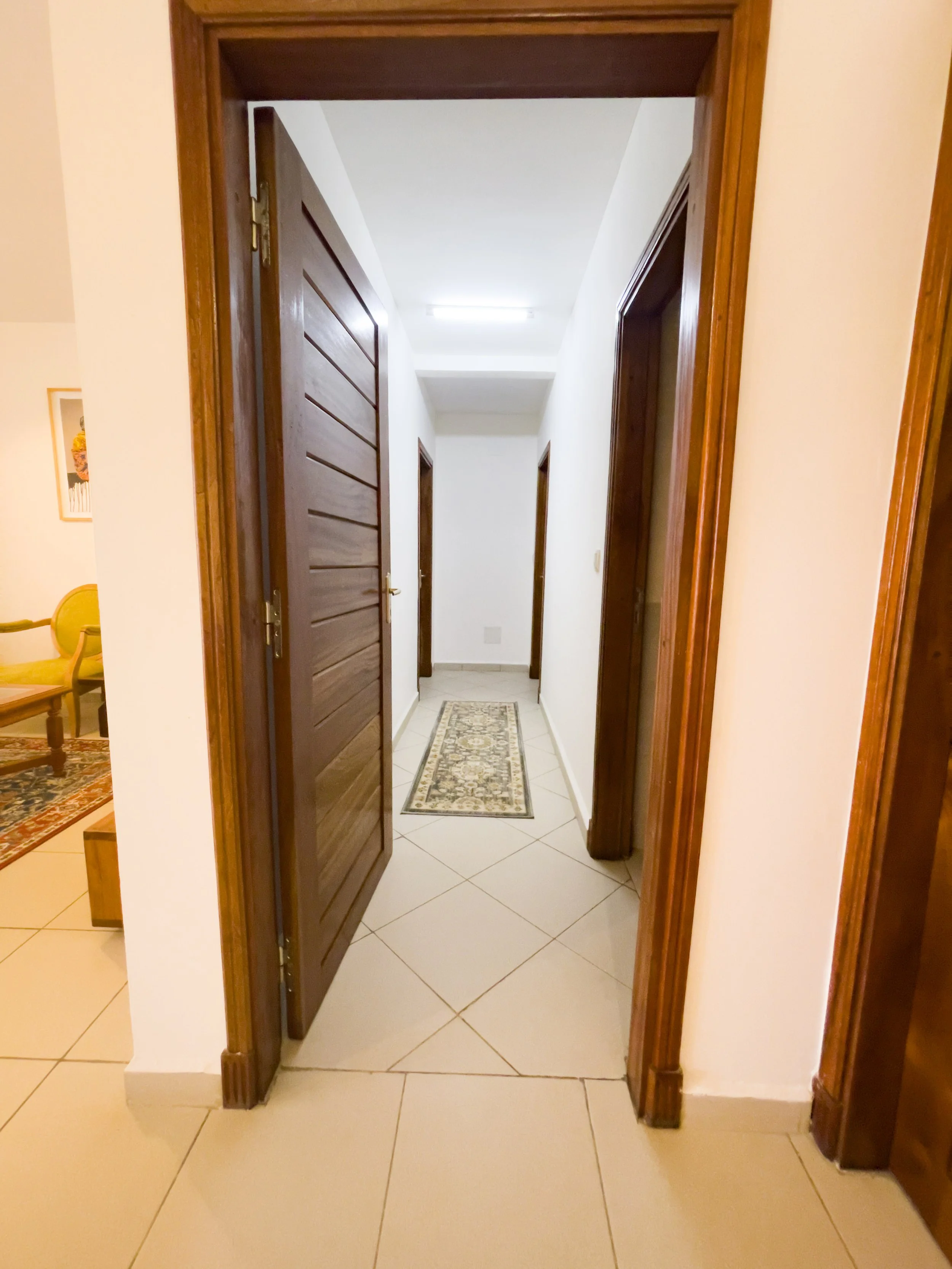 Vue d'un couloir intérieur avec portes en bois, tapis au sol, murs blancs et éclairage fluorescent au plafond.