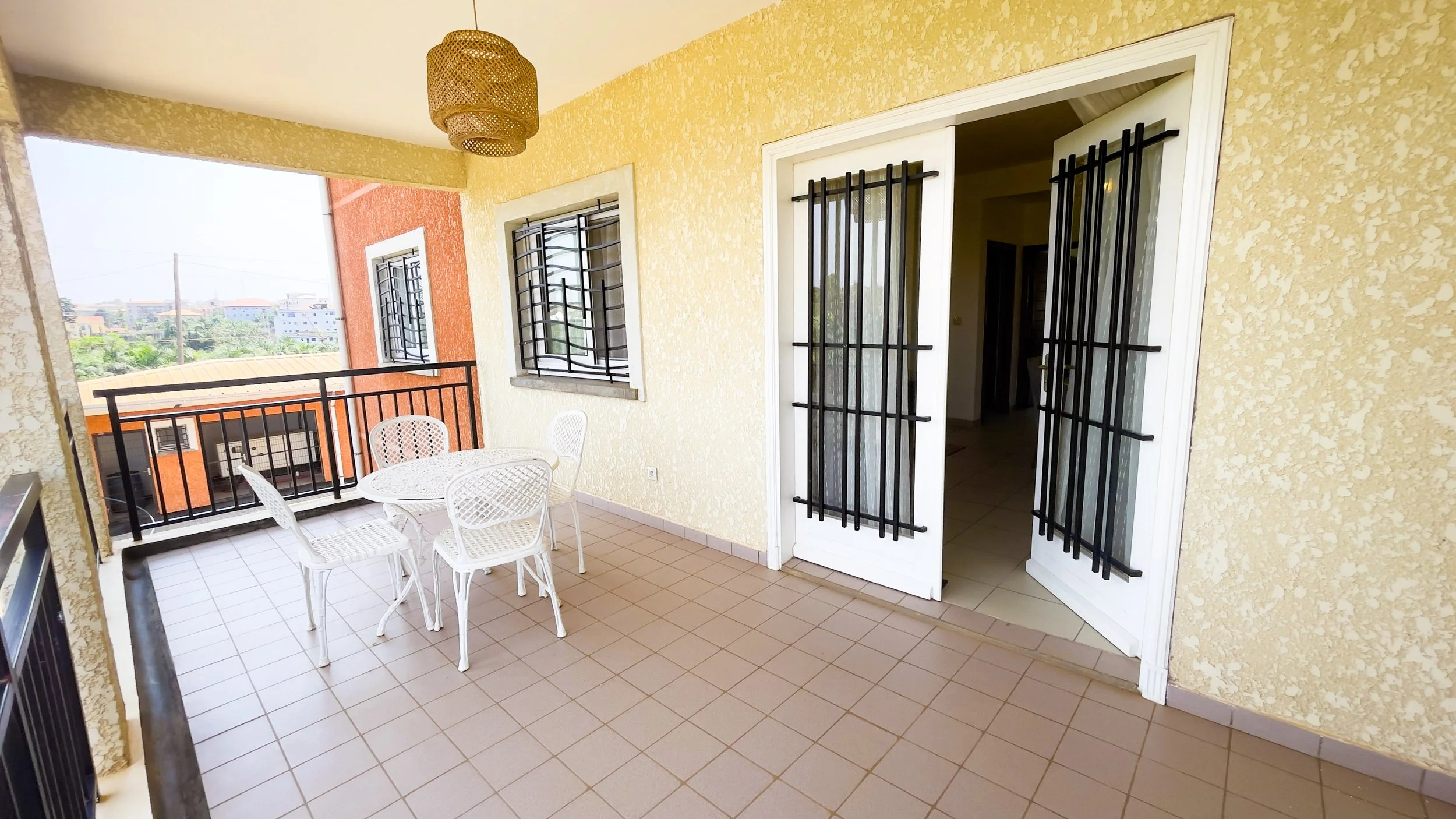 Balcon avec une table ronde en plastique blanc et des chaises, murs jaunes, fenêtres avec barreaux noirs, porte-fenêtre avec grille de sécurité, vue sur des bâtiments et un ciel clair.