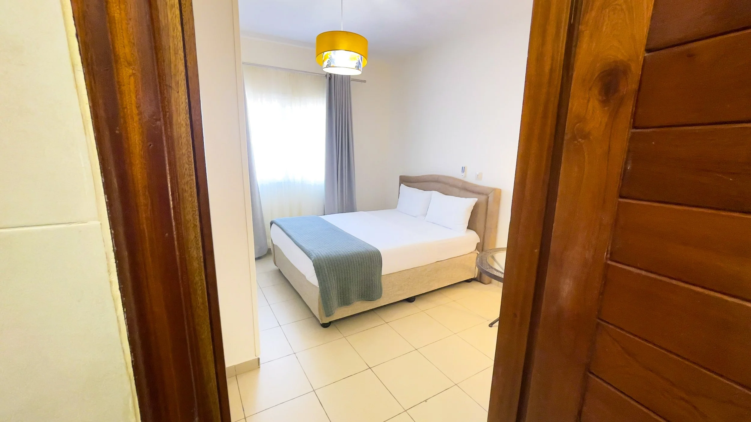 Chambre d'hôtel avec lit double, fenêtres avec rideaux, lampadaire jaune, côté bois de la porte d'entrée.