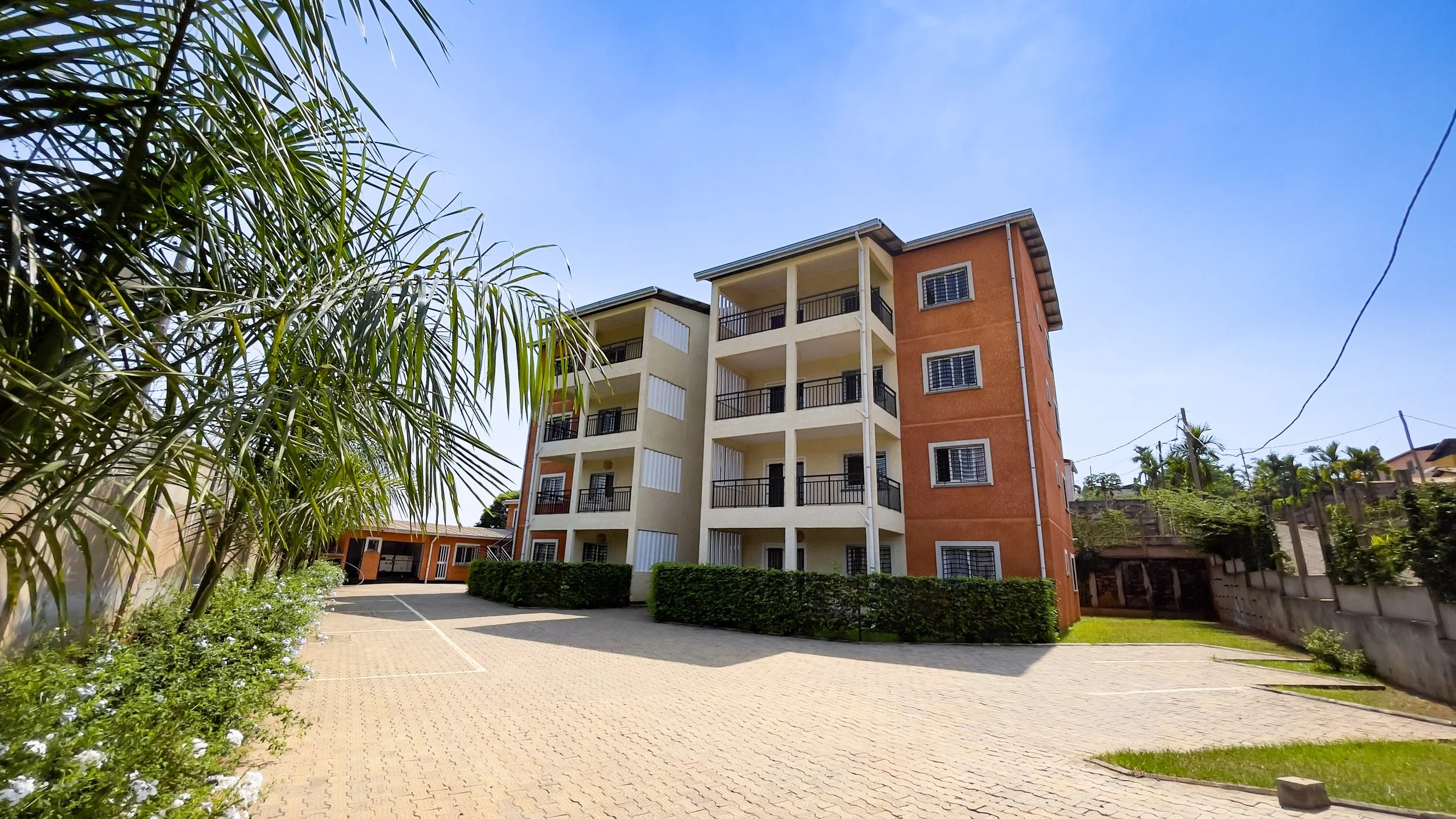 Immeuble résidentiel avec plusieurs étages, volets aux fenêtres, balcon avec garde-corps noirs, entouré de végétation, dans un parking pavé, sous un ciel bleu clair.