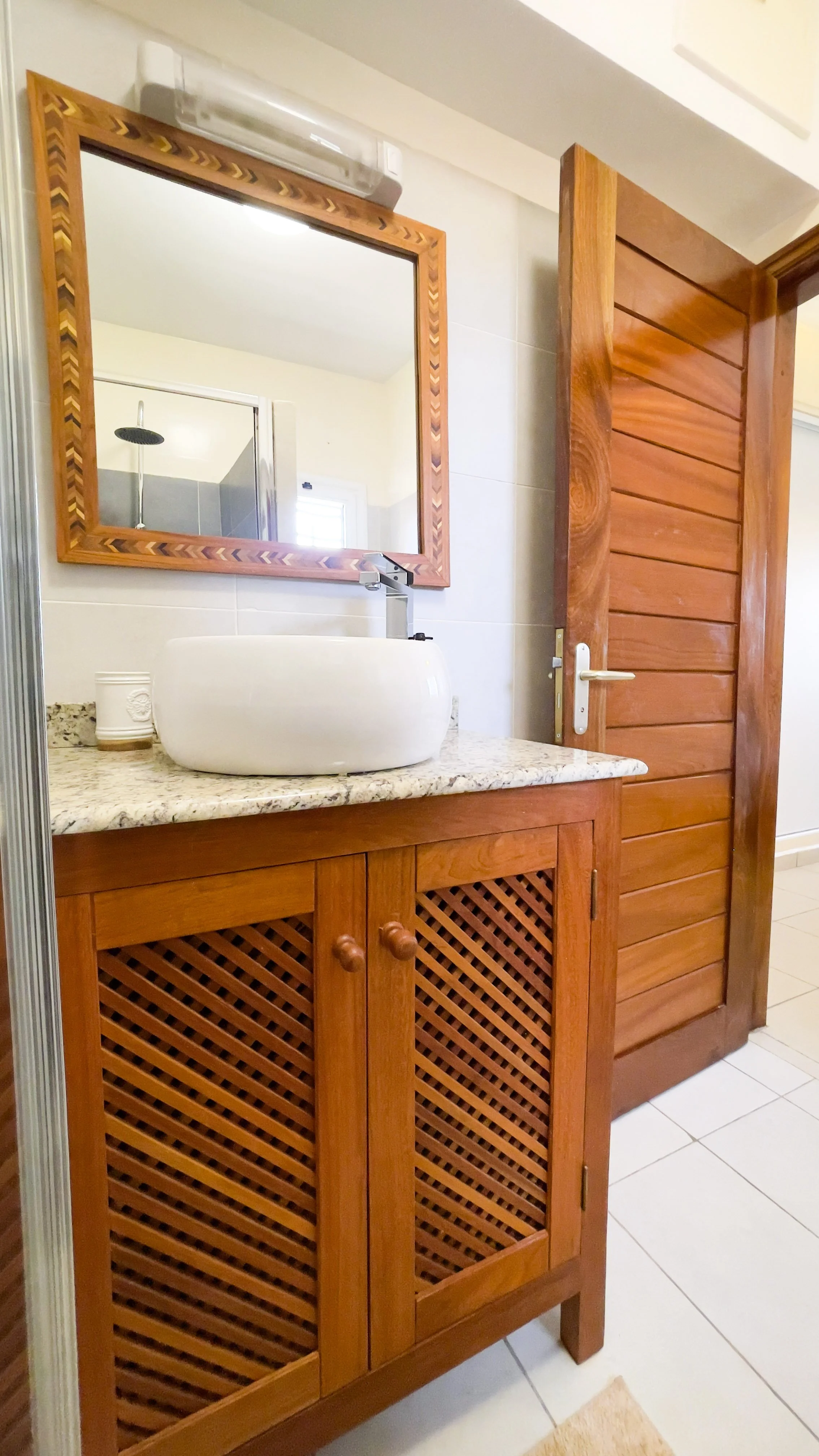 Salle de bain avec un meuble en bois avec un lavabo blanc rond sur un plan en granite, un miroir encadré en bois, une porte en bois, et un mur avec un miroir ou une fenêtre dans le reflet.