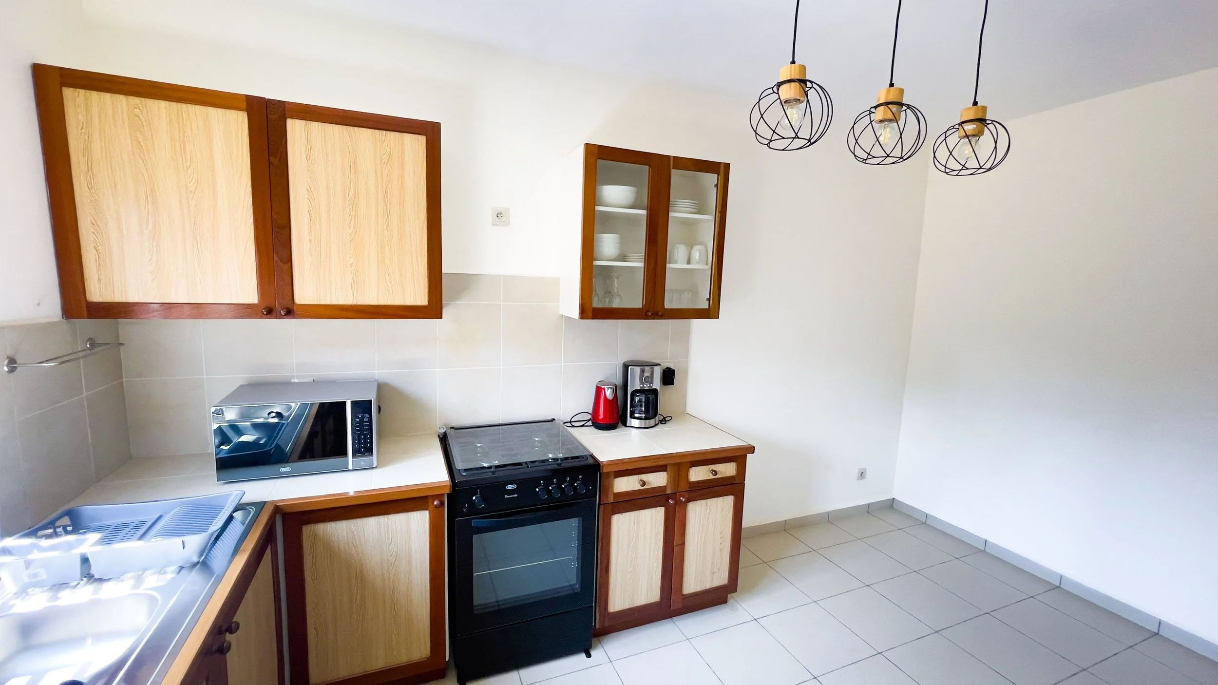 Cuisine simple avec placards en bois clair, micro-ondes, cuisinière électrique, cafetière, bouilloire, et évier.