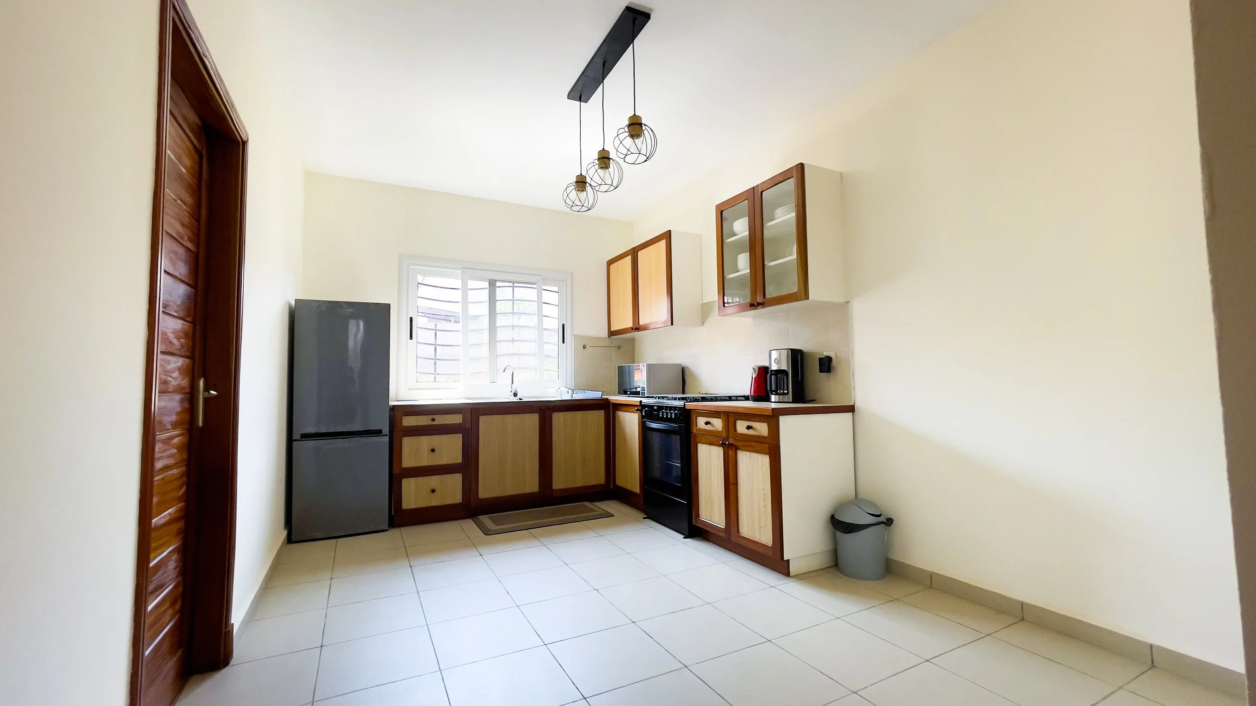 Cuisine simple avec des armoires en bois, un réfrigérateur gris, un four, une cafetière, et une fenêtre avec volets, carrelage clair au sol, suspendu avec un luminaire moderne.