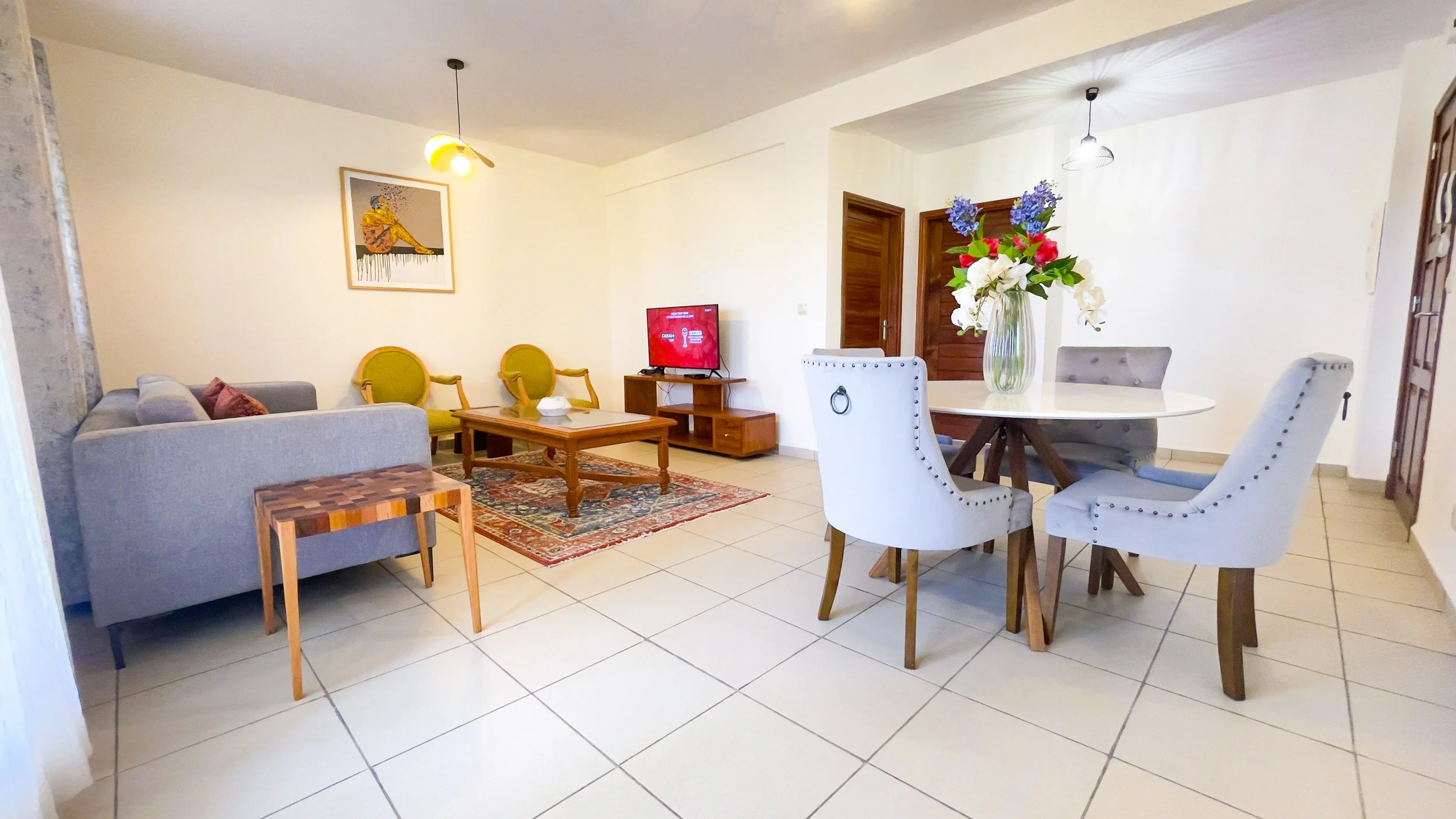 Salon avec canapé gris, fauteuils verts, table basse, tapis, télévision, salle à manger avec vase de fleurs, murs blancs, sol en carreaux