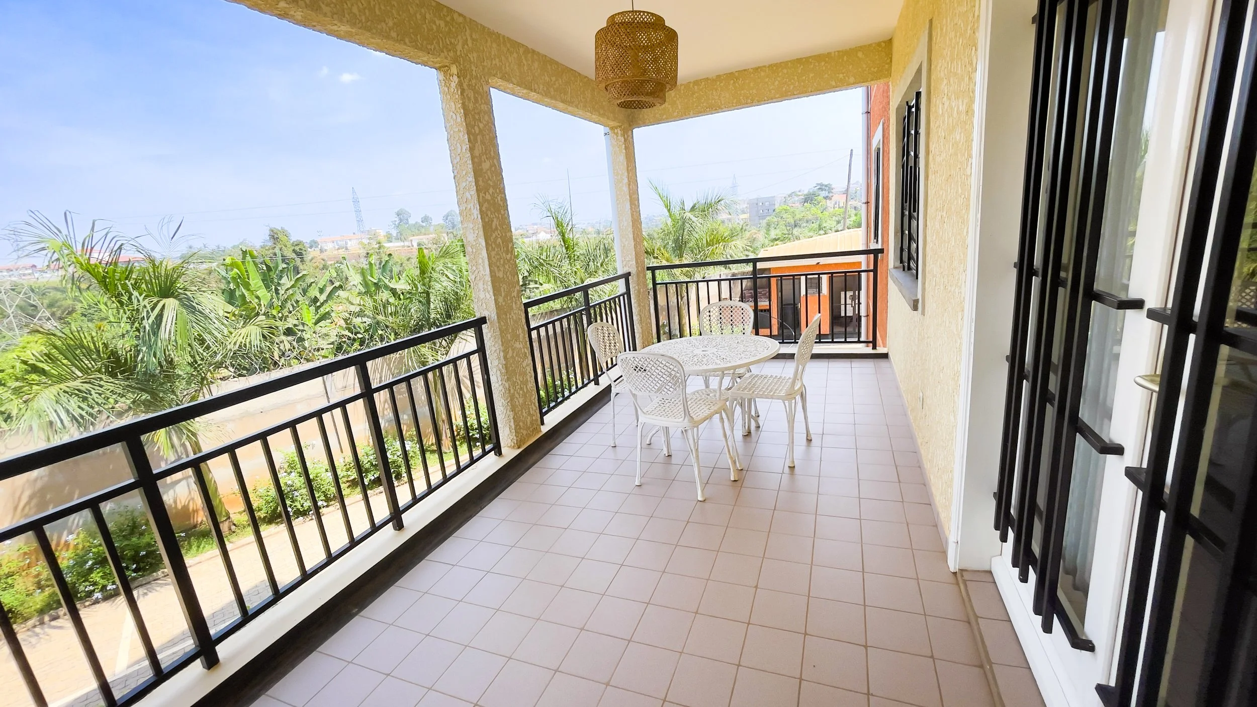 Balcon avec une table ronde et quatre chaises blanches, entouré d'une rambarde noire, vue sur un paysage verdoyant avec des arbres et un ciel clair.
