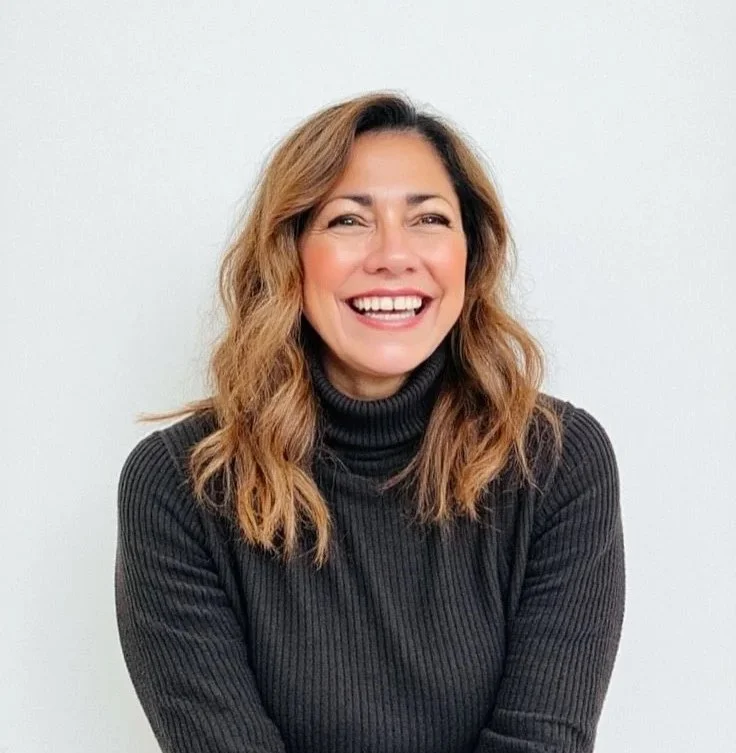 A woman with shoulder-length wavy light brown hair smiling, wearing a black ribbed turtleneck sweater, standing against a plain white background.