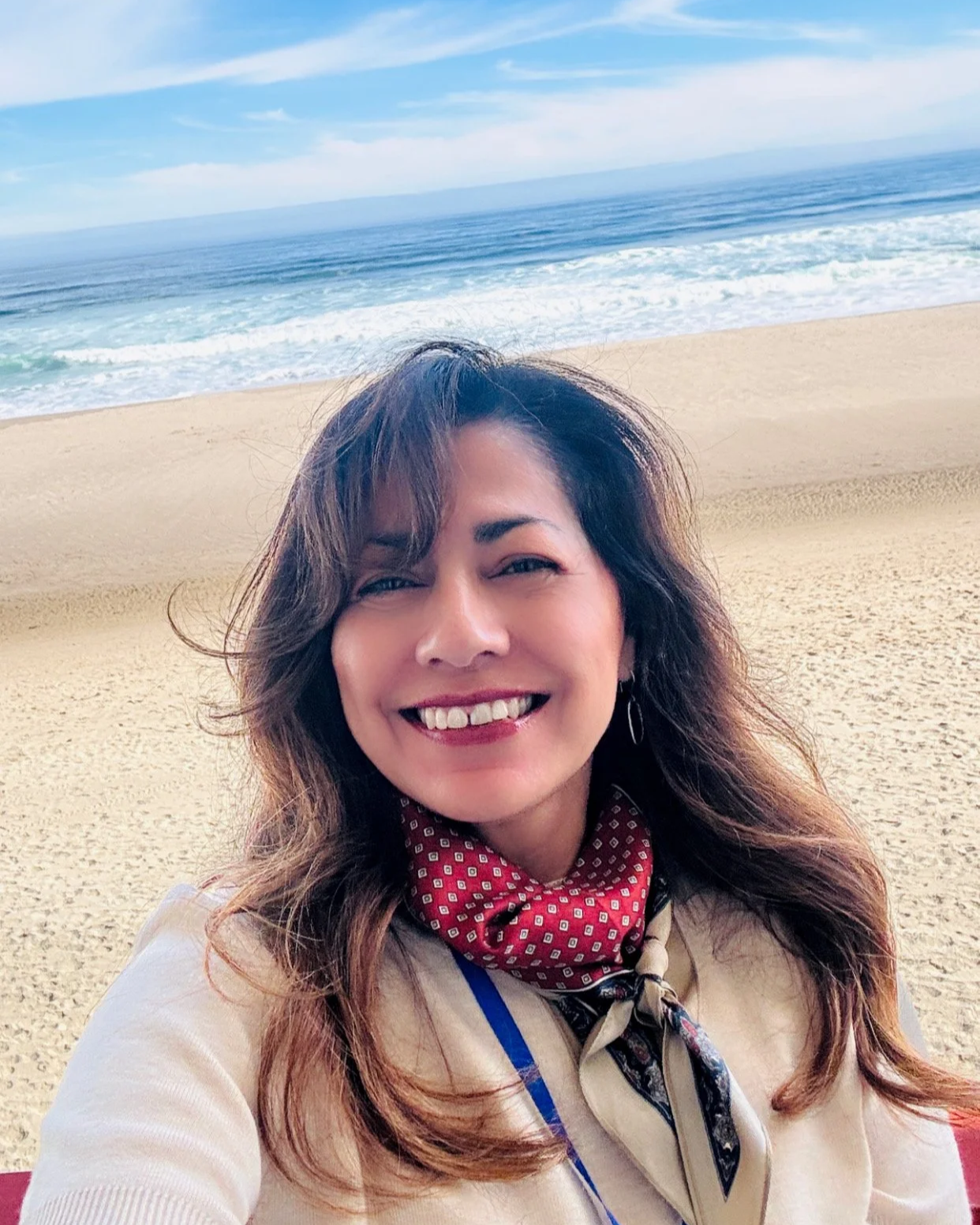 A woman smiling at the camera on a sandy beach with the ocean and blue sky in the background.