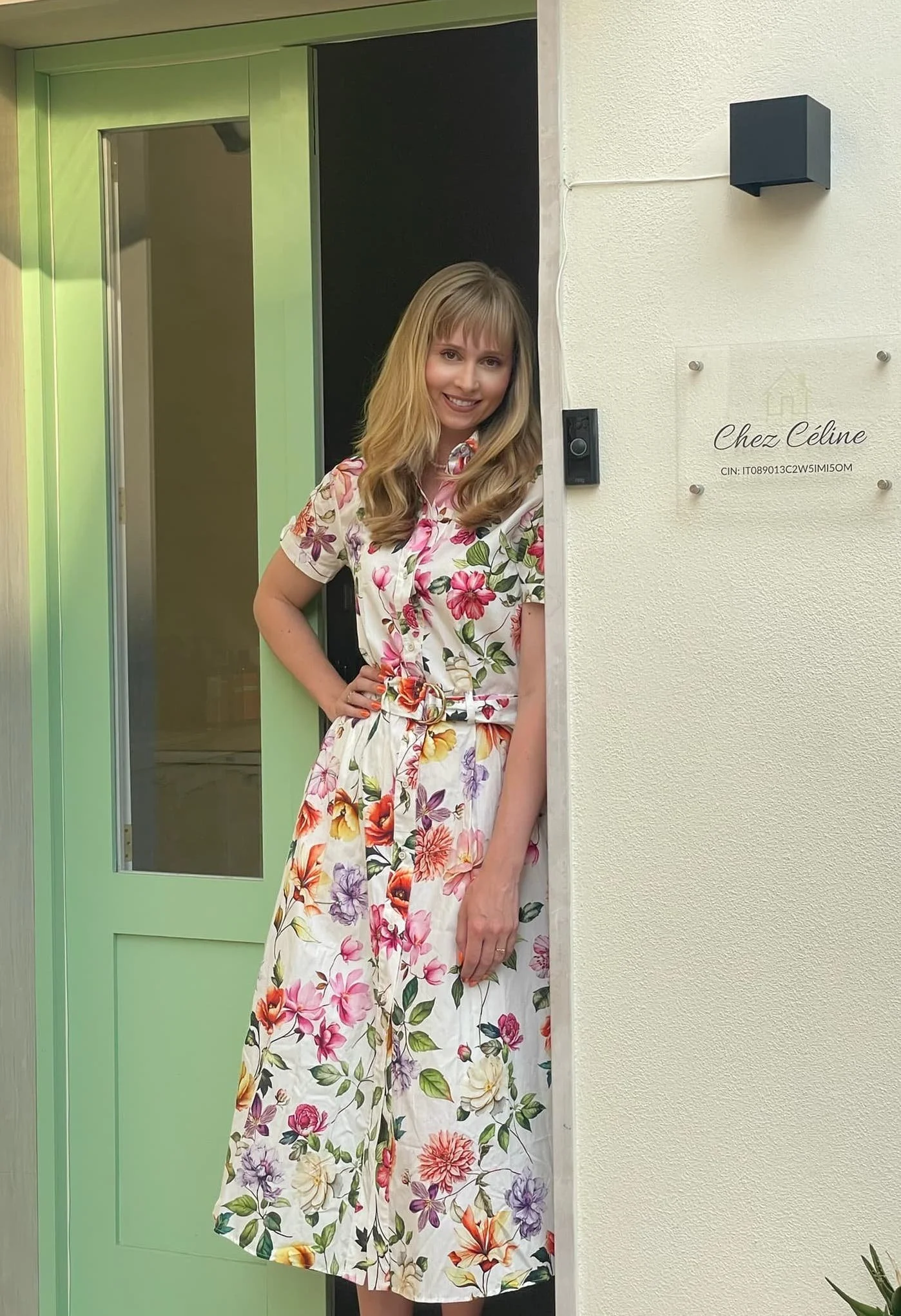 A woman with blonde hair wearing a floral dress standing at the door of a house, smiling.