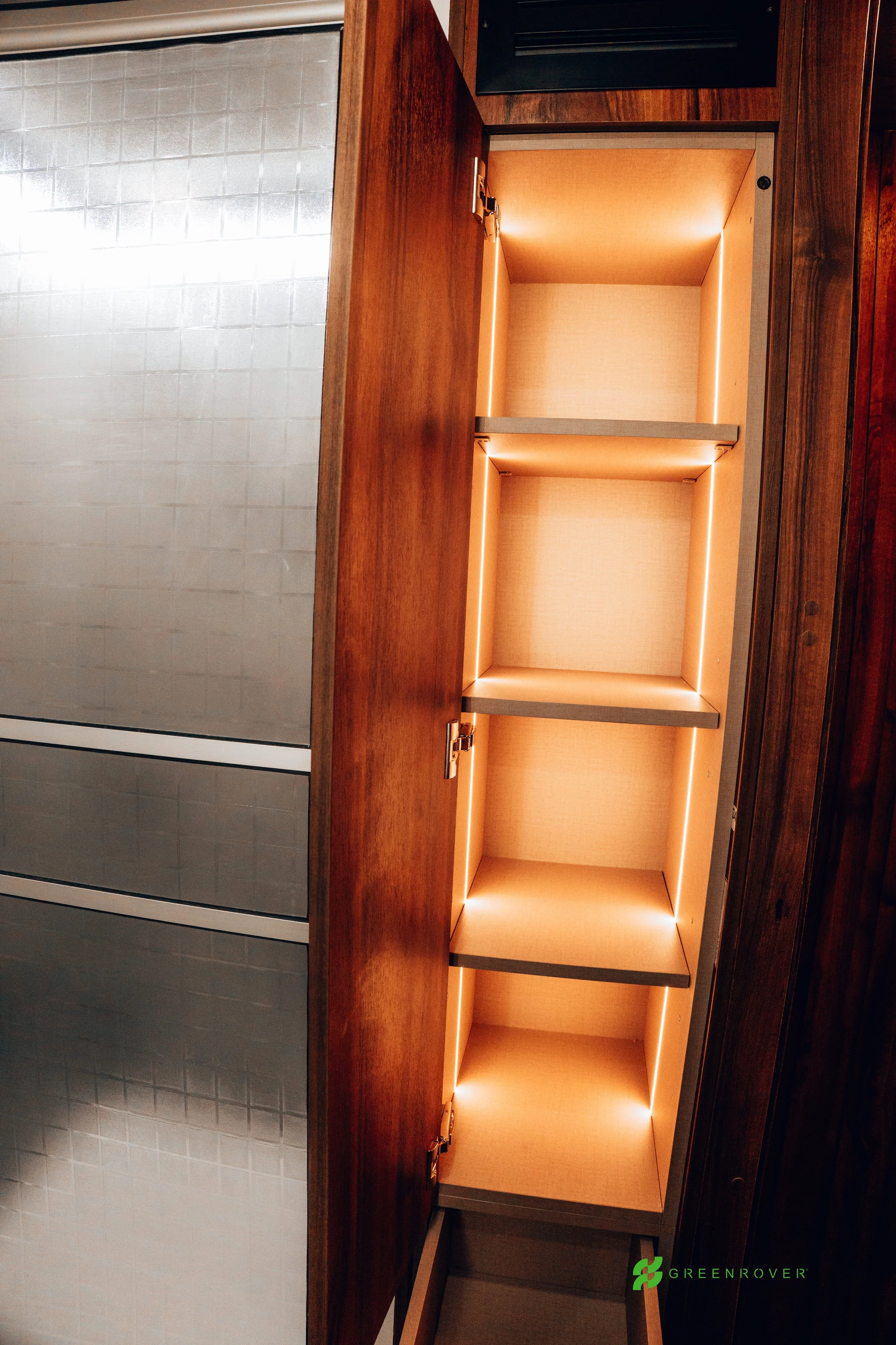 Open wooden cabinet with illuminated interior and three shelves, next to tiled wall in a room.