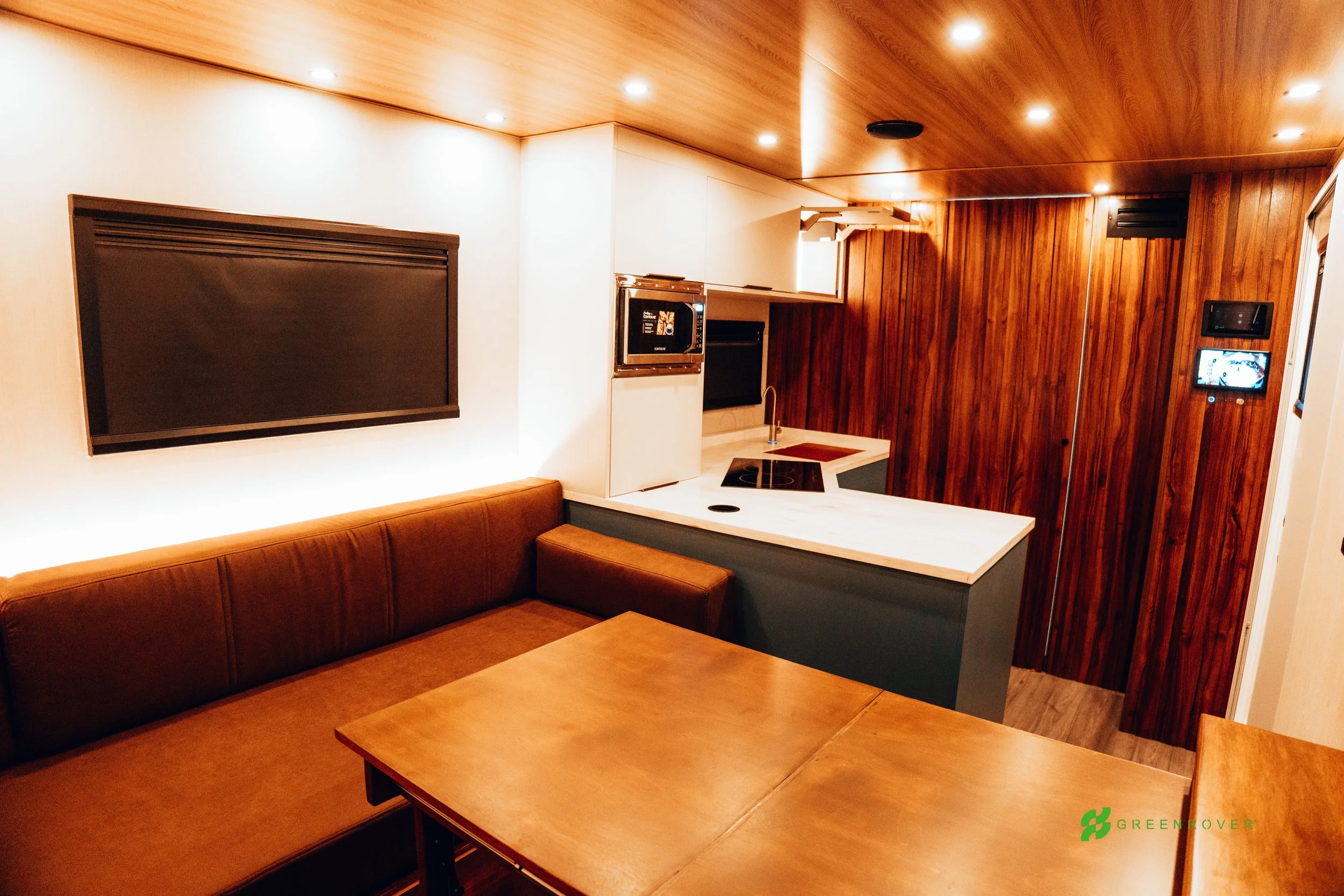 Interior of a modern kitchen and dining area with wood paneling ceiling and wall, a brown leather banquette, a wooden dining table, a white kitchen counter with an electric stove, small sink, microwave, and a small TV on the wall.