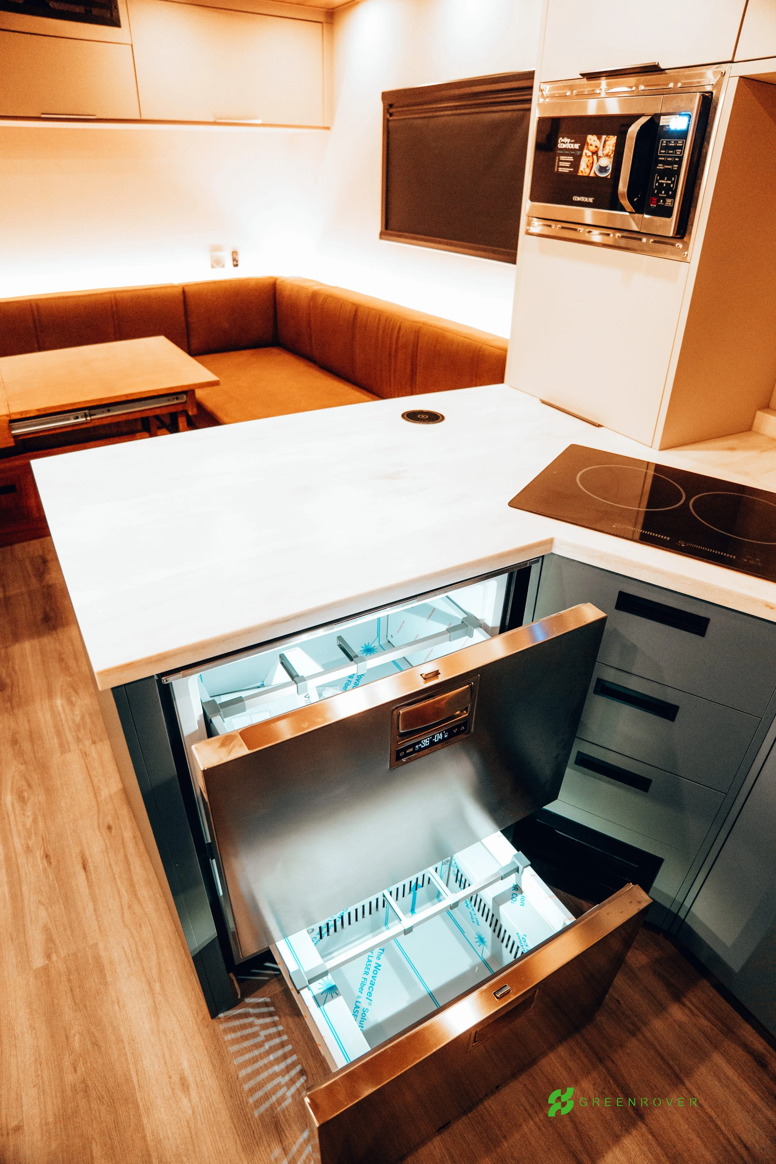 Modern kitchen with an open freezer drawer, a white marble countertop, a built-in microwave, a black induction cooktop, and a brown upholstered corner sofa in the dining area.