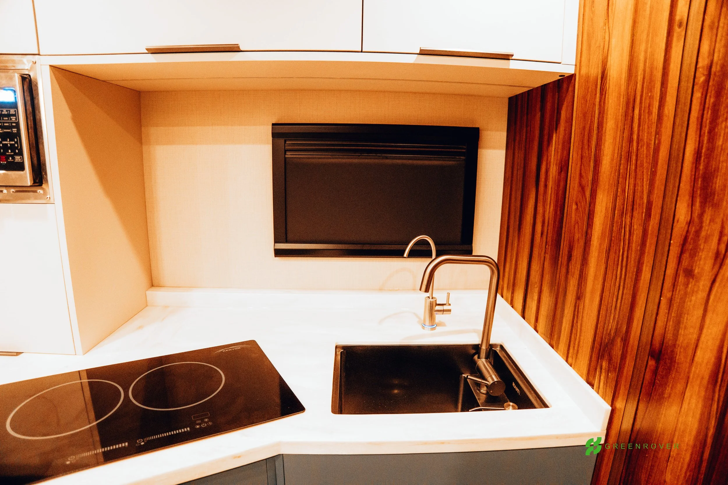 Kitchen counter with a black electric stove, a sink with a faucet, a small black appliance, and a wood-paneled wall, with a beige backsplash.