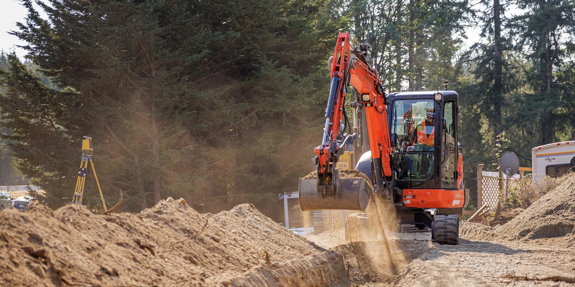 Construction site with an orange excavator digging into dirt, with a background of trees and construction equipment.