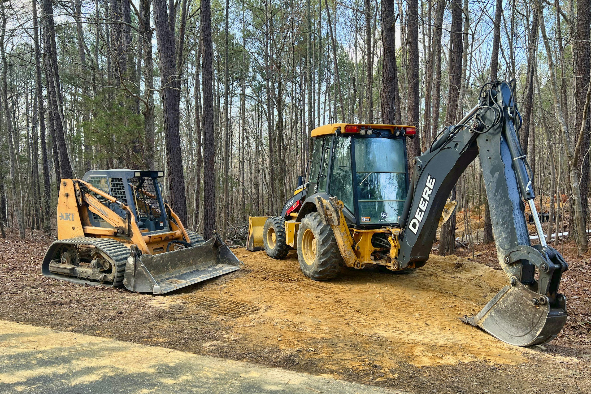 Construction site in a wooded area with two pieces of heavy machinery: a small track loader on the left and a mini excavator on the right, working on leveling earth.