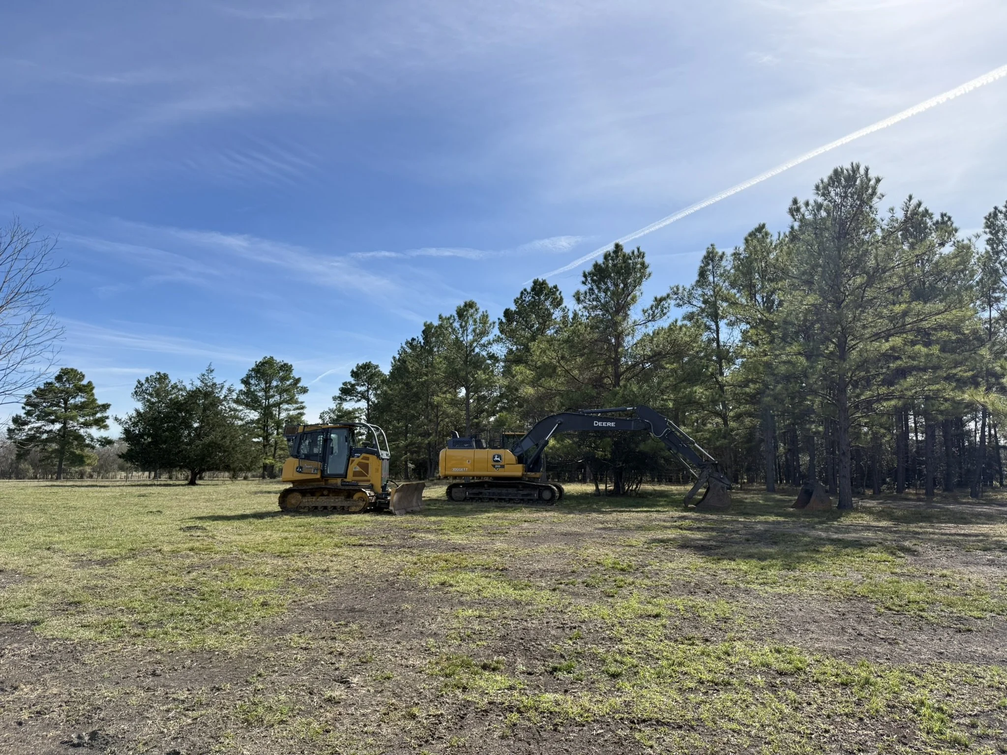 A wide view of a construction site in a grassy field with two yellow and black excavators parked beside trees under a blue sky with wispy clouds.